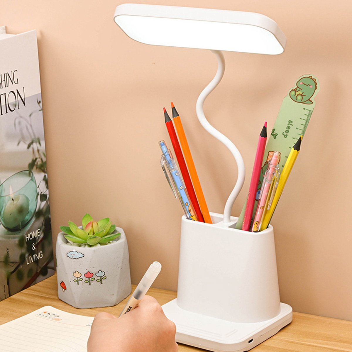 Ein Schreibtisch mit Lampe, Bleistiften und einer Sukkulente. Eine Hand schreibt in ein Notizbuch. Hintergrund ist eine Wand und ein Buch.