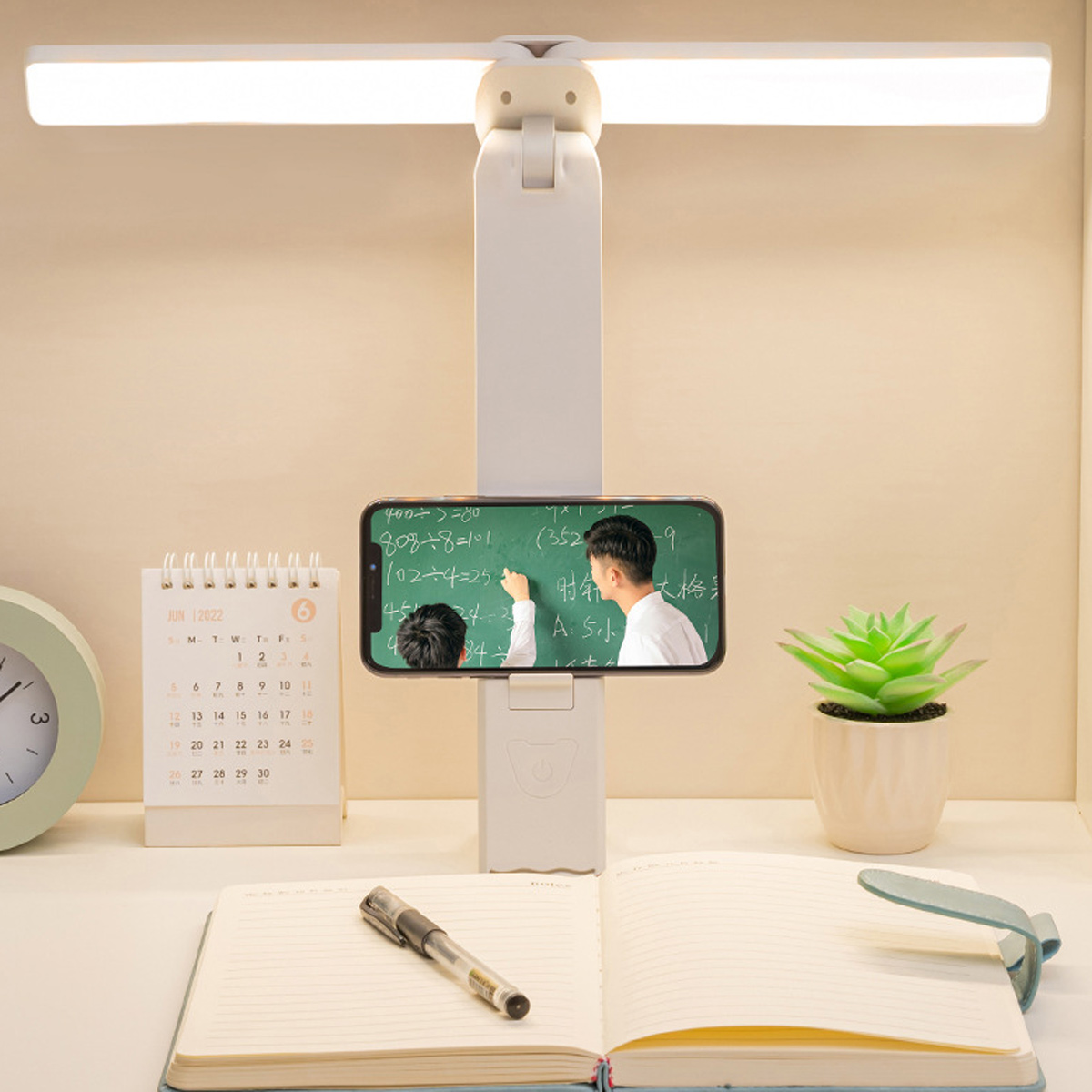 Ein Schreibtisch mit Lampe, Telefon mit Mathe-Lektion, Kalender, Pflanze, Notizbuch und Stift. Das Telefon zeigt eine Person, die auf eine Tafel schreibt.