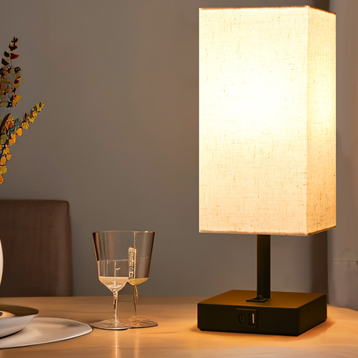 Eine beleuchtete rechteckige Lampe auf schwarzem Sockel steht auf einem Tisch. Zwei Weingläser stehen links. Weiches Licht und neutraler Hintergrund.