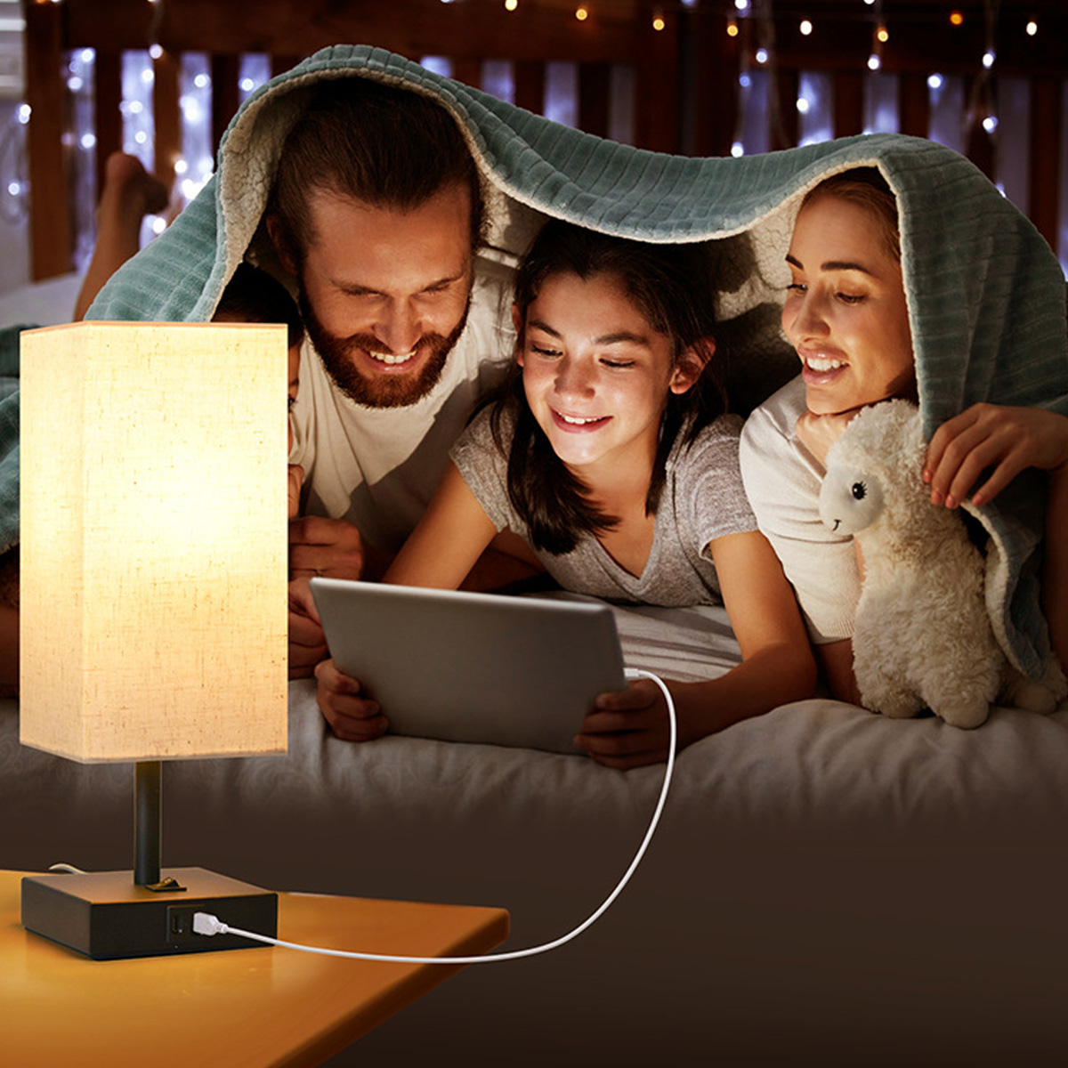 Familie unter einer Decke, schaut auf ein Tablet. Eine Lampe steht daneben. Ein Stofftier wird gehalten. Im Hintergrund Lichterketten.