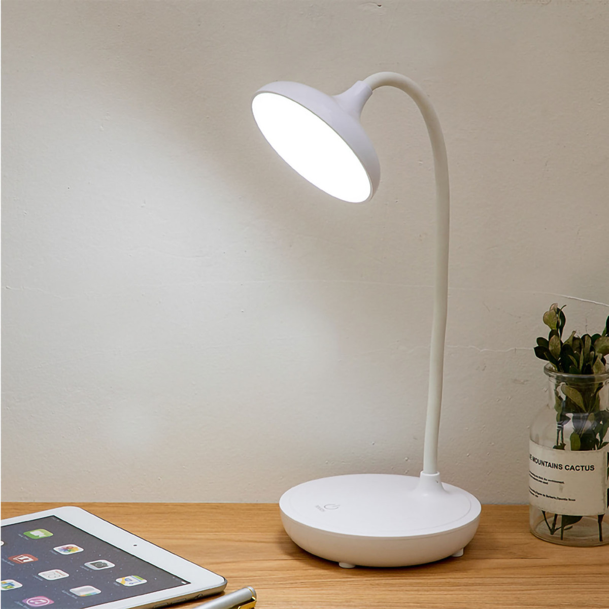 Eine weiße Schreibtischlampe steht auf einem Holztisch. Ein Tablet, ein Stift und eine Pflanze in einem Glas sind ebenfalls zu sehen.