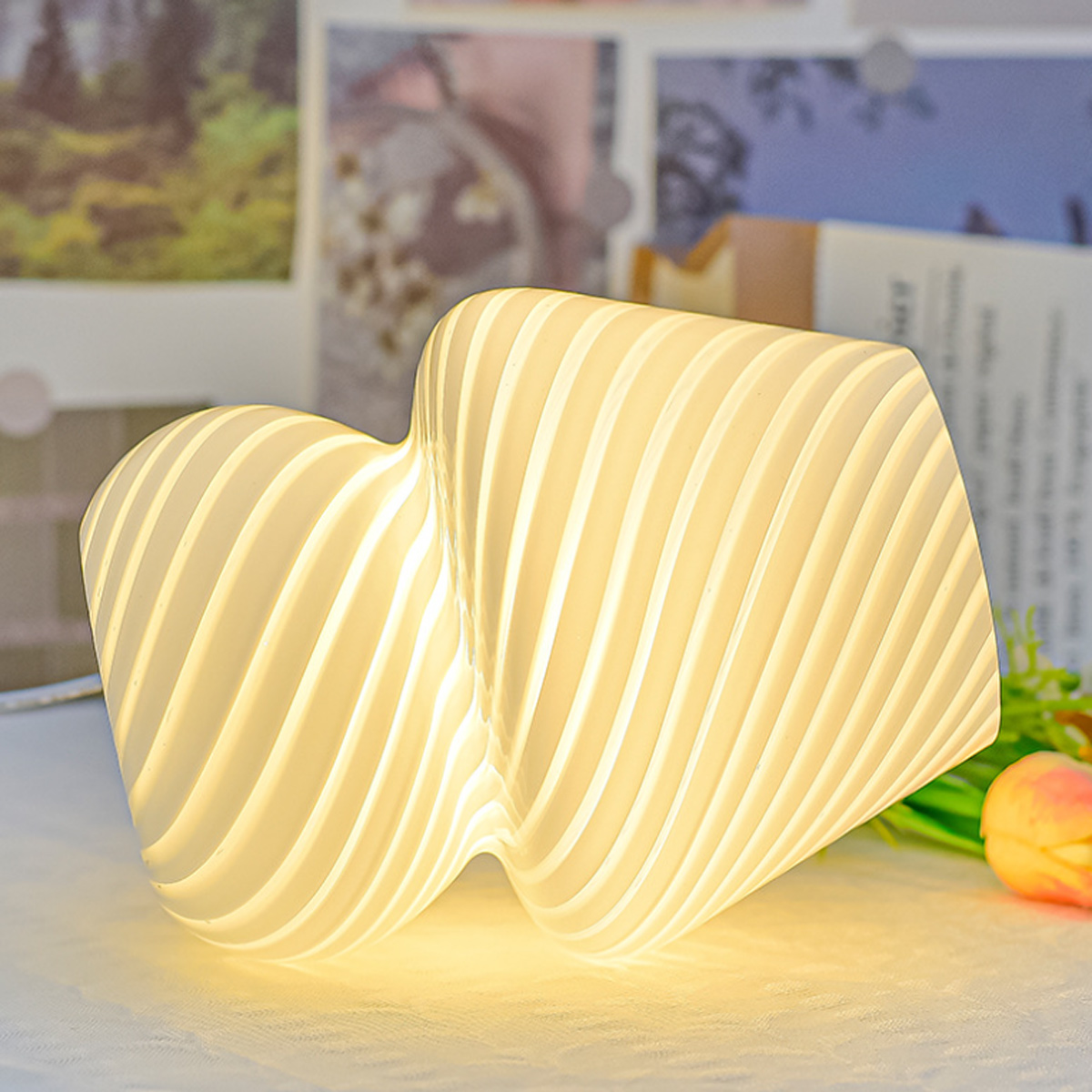 Eine weiße, wellenförmige Lampe strahlt ein sanftes Licht aus. Im Hintergrund verschwommen Bilder und eine Blume.