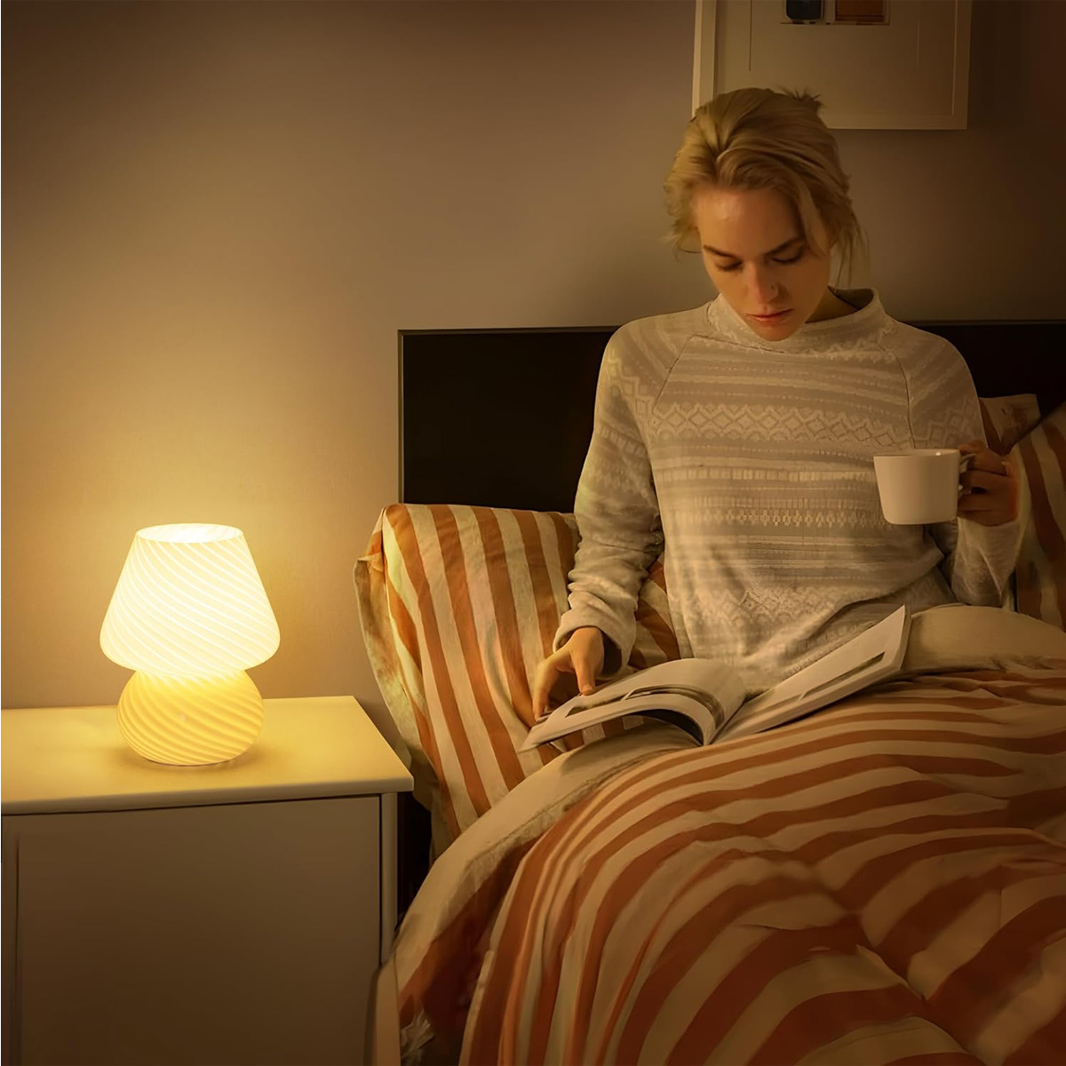 Frau im Bett liest mit einer Tasse. Eine Nachttischlampe ist an. Orange-weiß gestreifte Bettwäsche ist sichtbar.