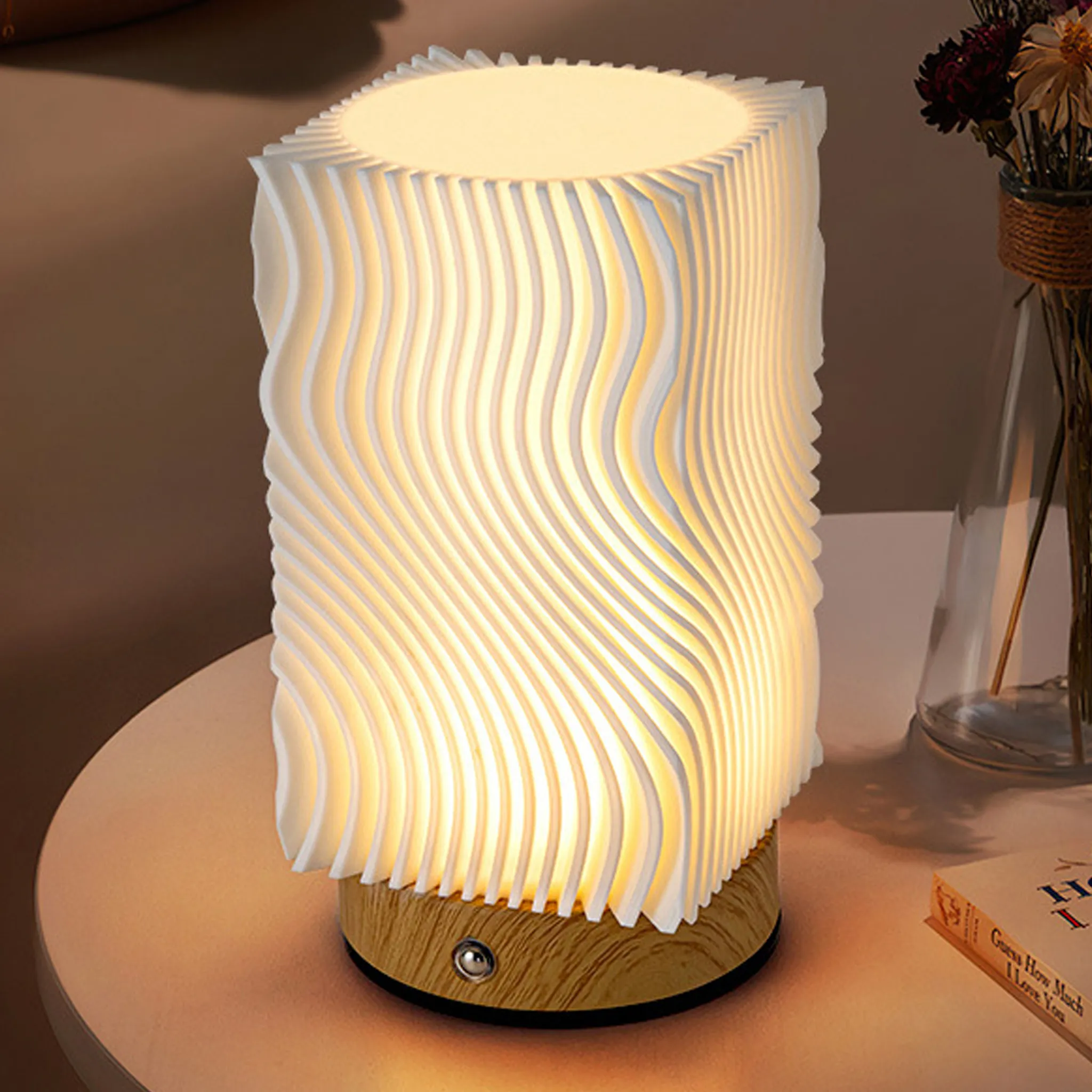 Eine beleuchtete, weiße, wellenförmige Lampe steht auf einem Tisch. Im Hintergrund sind eine Blumenvase und ein Buch zu sehen.