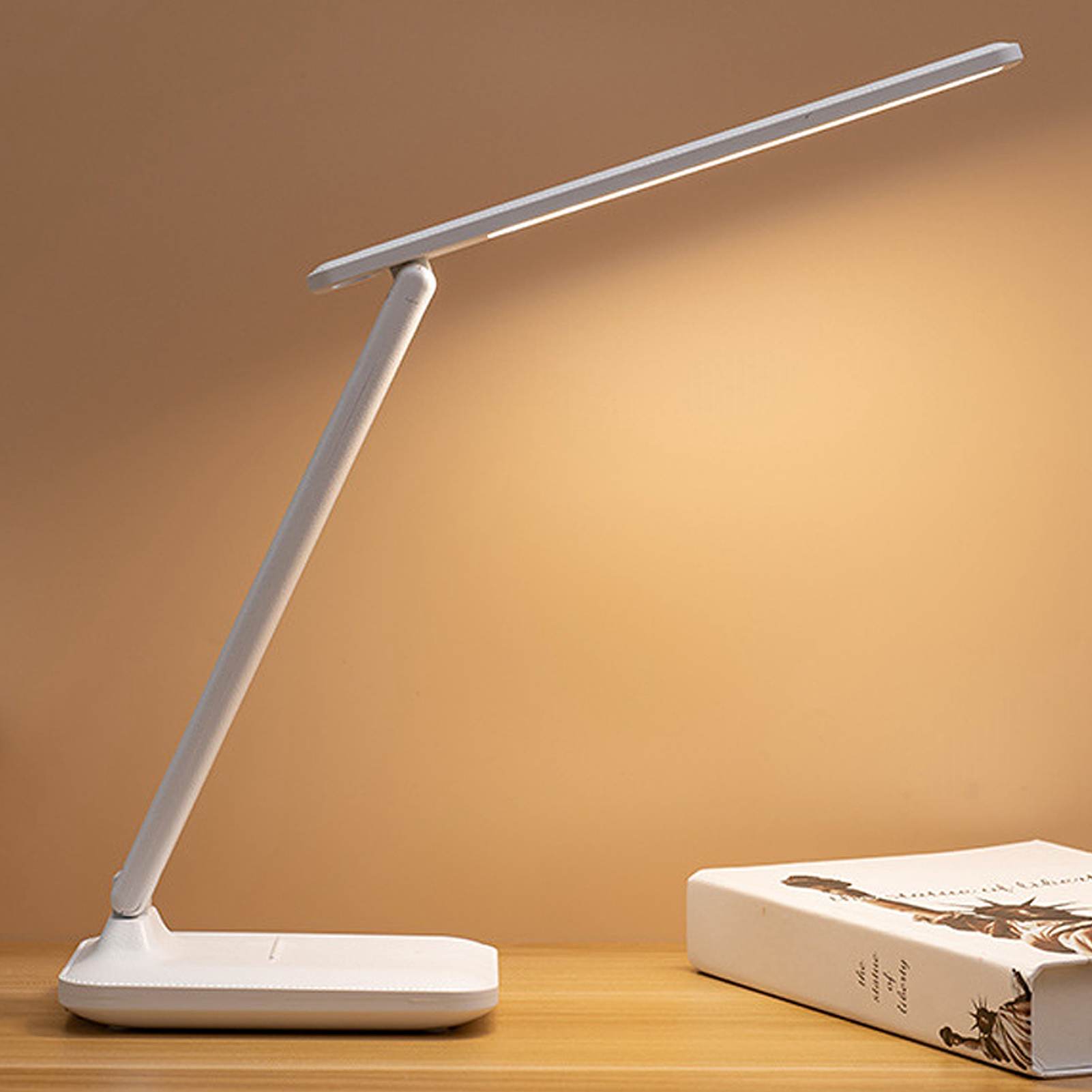 Eine weiße Schreibtischlampe leuchtet auf einem Holztisch mit einem Buch. Der Hintergrund ist eine hellbraune Wand.