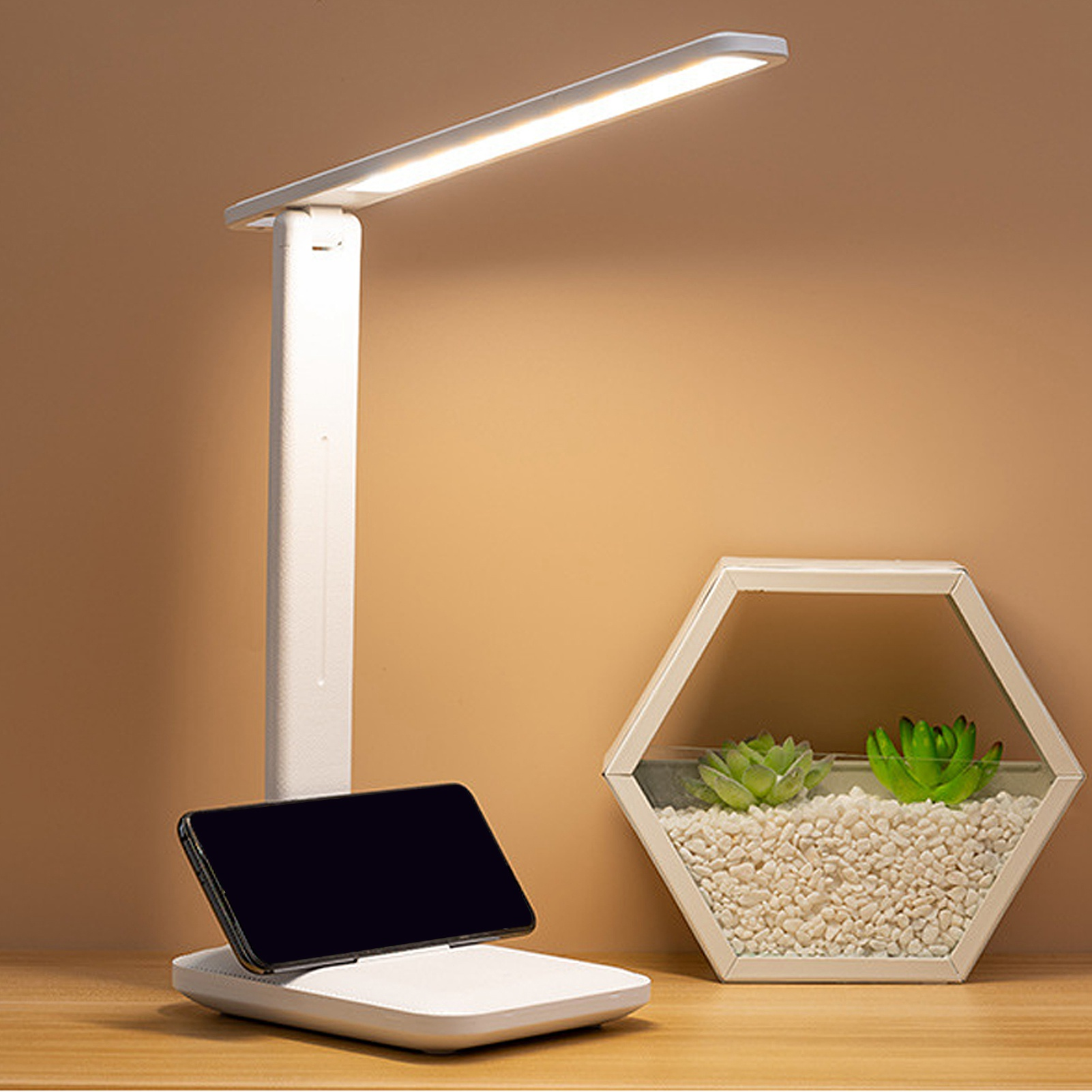 Eine weiße Schreibtischlampe beleuchtet ein Telefon auf einem Ladegerät. Daneben steht ein Terrarium mit Sukkulenten.