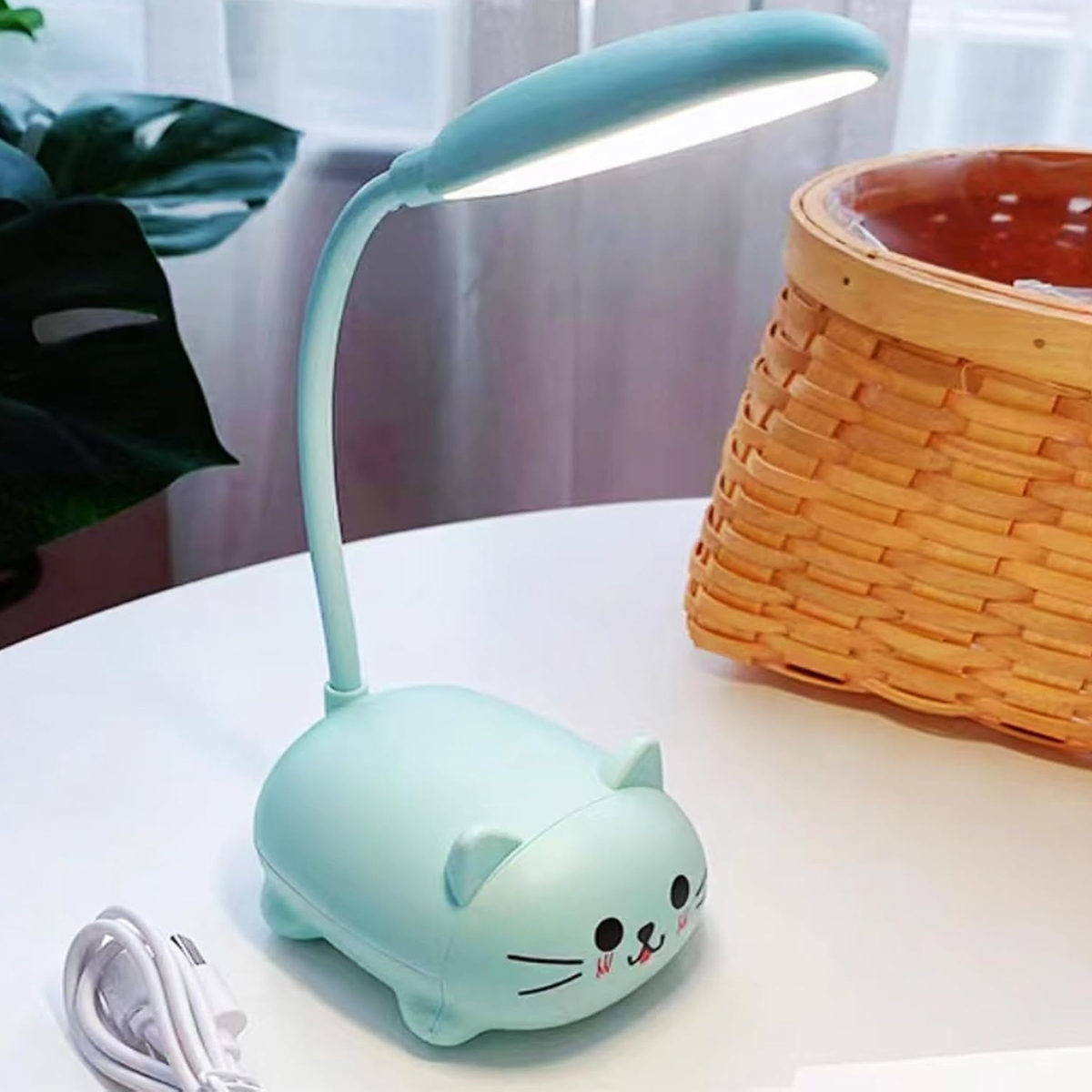 Eine mintgrüne, katzenförmige Schreibtischlampe mit flexiblem Hals und beleuchtetem Lampenkopf, neben einem Weidenkorb.