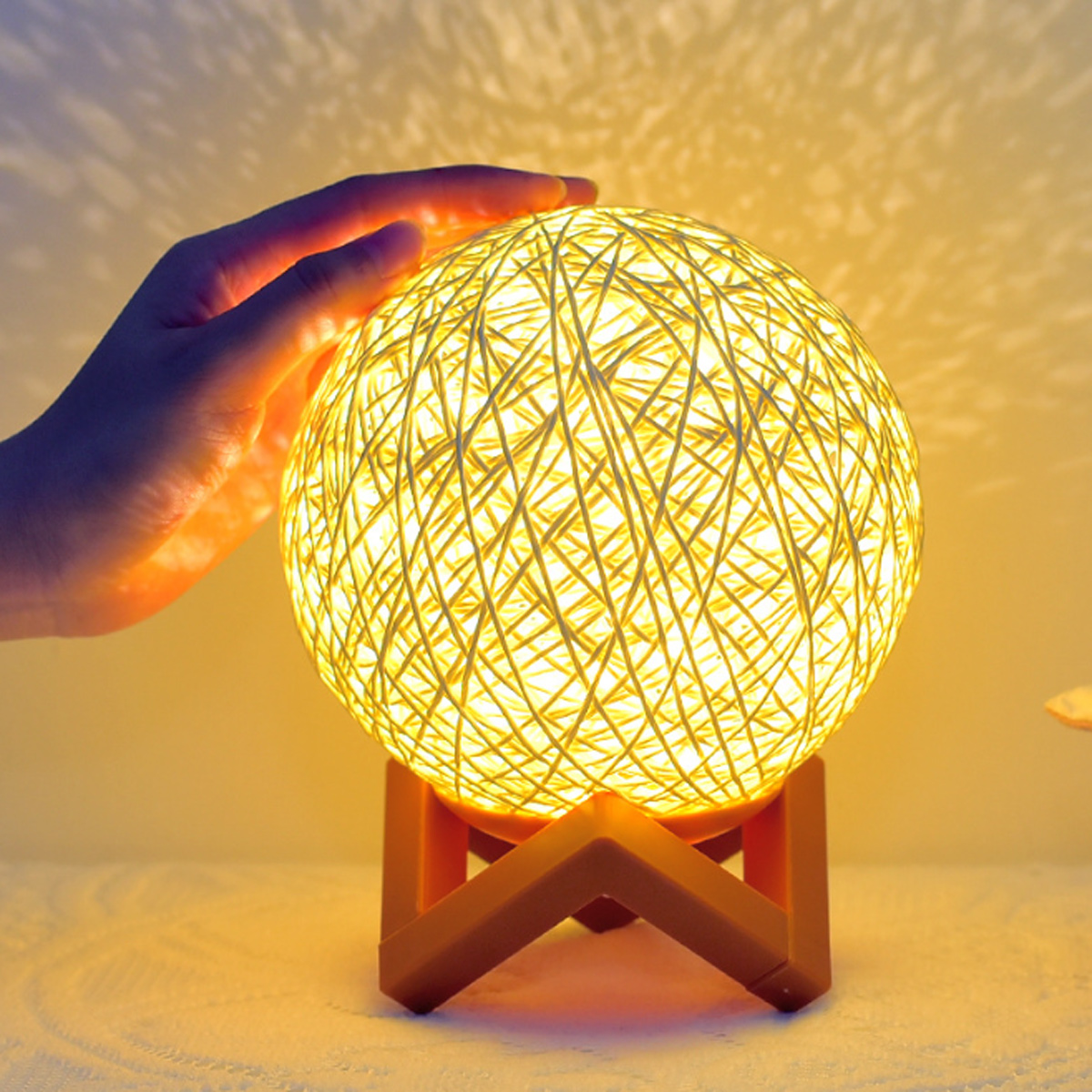 Eine Hand berührt eine leuchtende, kugelförmige Lampe aus gewebtem Material. Die Lampe steht auf einem Holzständer und wirft Licht.