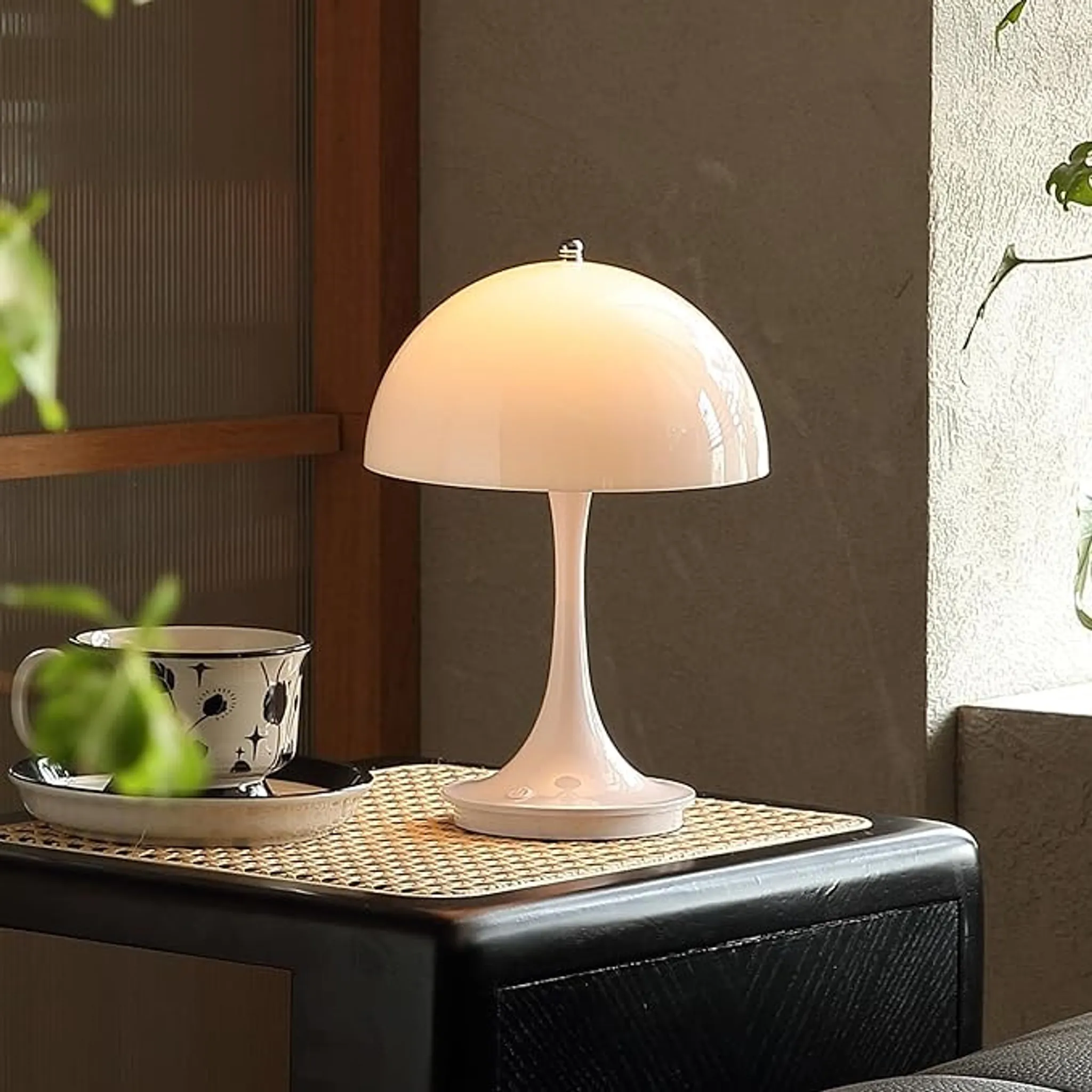 Eine weiße Pilzlampe steht auf einem schwarzen Tisch mit einer Tasse und Untertasse. Der Hintergrund ist eine Wand und ein Fenster.