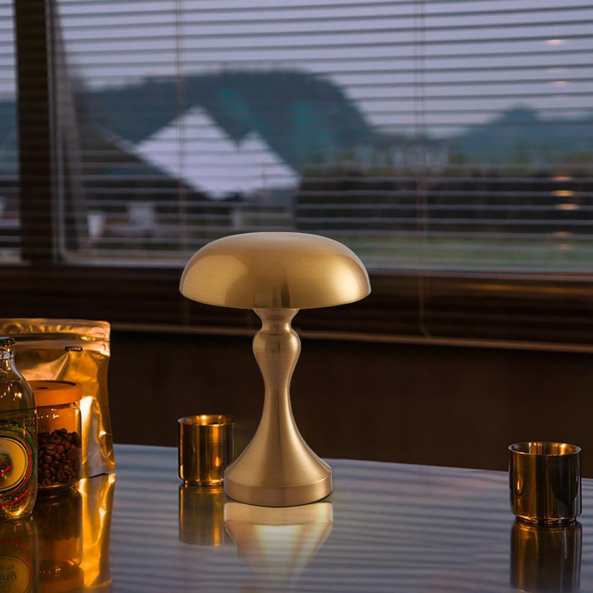 Eine goldene Pilzlampe steht auf einer reflektierenden Oberfläche. Dahinter ein Fenster mit Jalousien und verschwommenem Bergblick.
