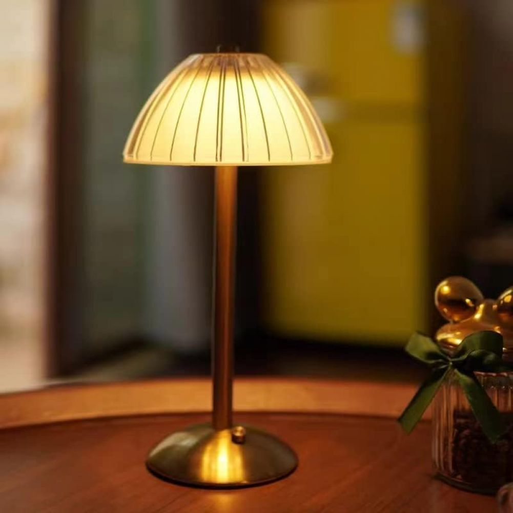 Eine beleuchtete Tischlampe mit Lampenschirm. Es steht auf einem Holztisch. Rechts steht ein Glas mit grünem Band.