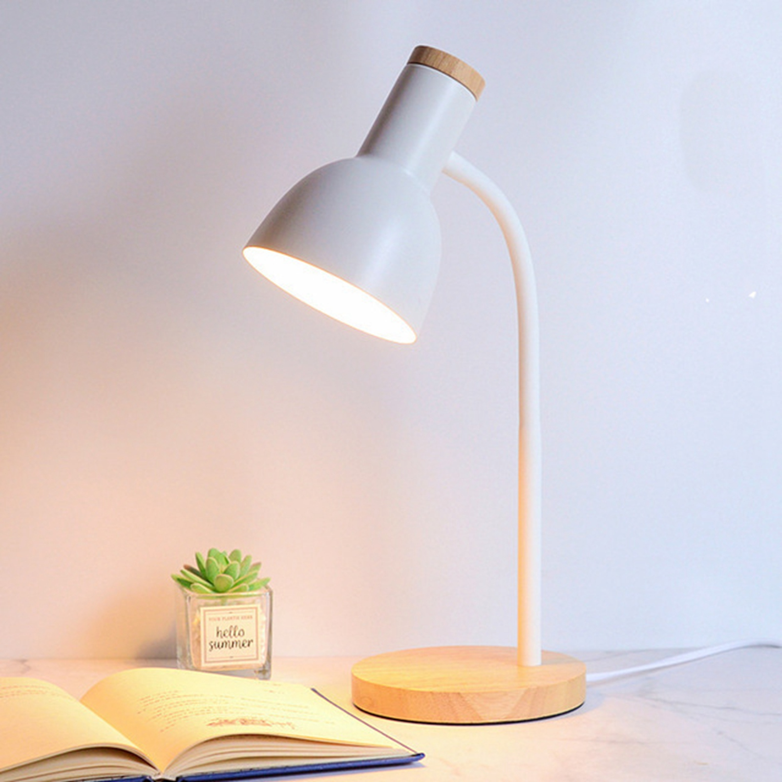 Eine weiße Schreibtischlampe mit Holzsockel beleuchtet einen Schreibtisch. Eine kleine Pflanze und ein offenes Buch liegen auf dem Tisch.