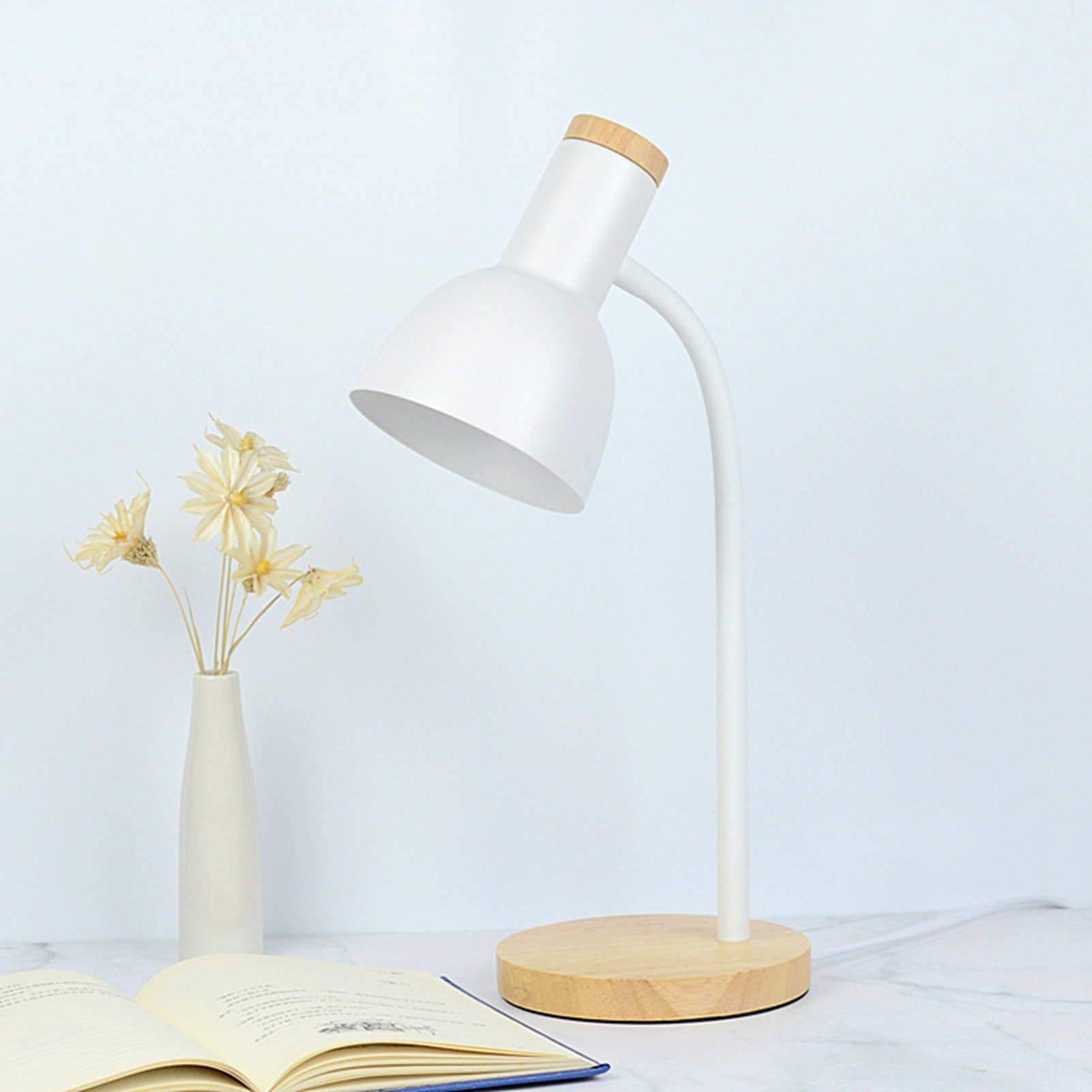 Eine weiße Schreibtischlampe mit Holzsockel, eine Vase mit Blumen und ein offenes Buch auf weißer Oberfläche.