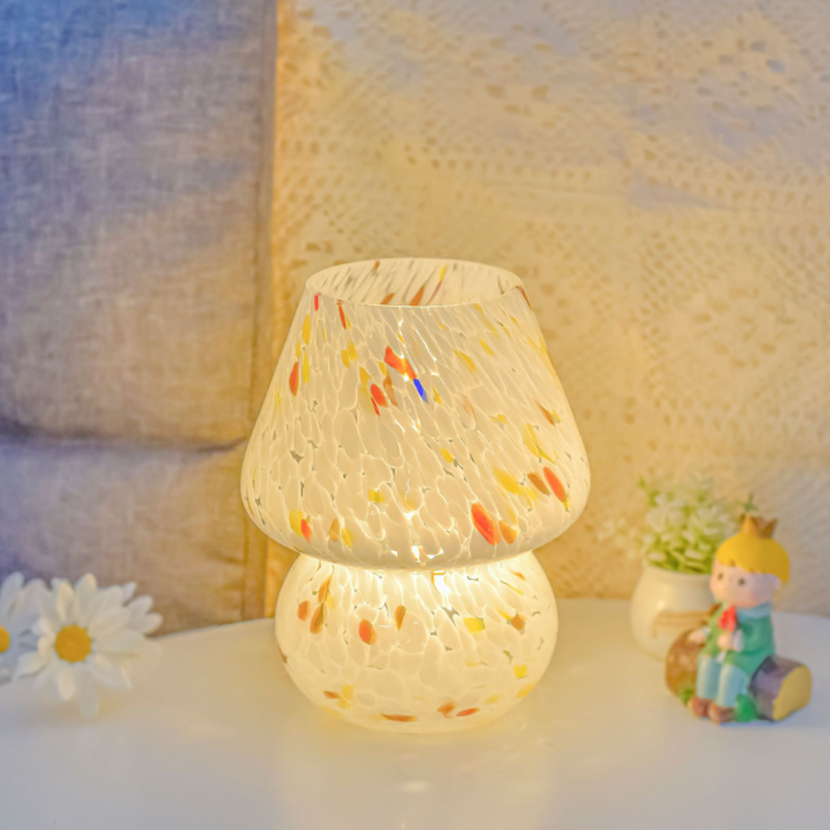 Eine pilzförmige Lampe mit bunten Sprenkeln steht auf einem weißen Tisch. Eine Statue und Blumen sind im Hintergrund.