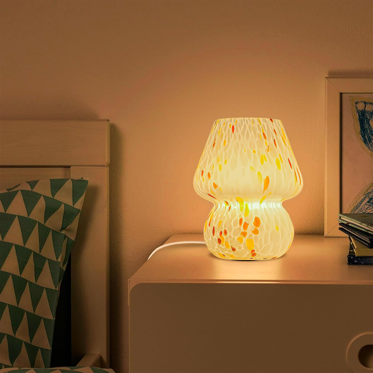 Eine pilzförmige Lampe mit bunten Punkten steht auf einem Nachttisch. Ein Bett und ein Bilderrahmen sind im Hintergrund.
