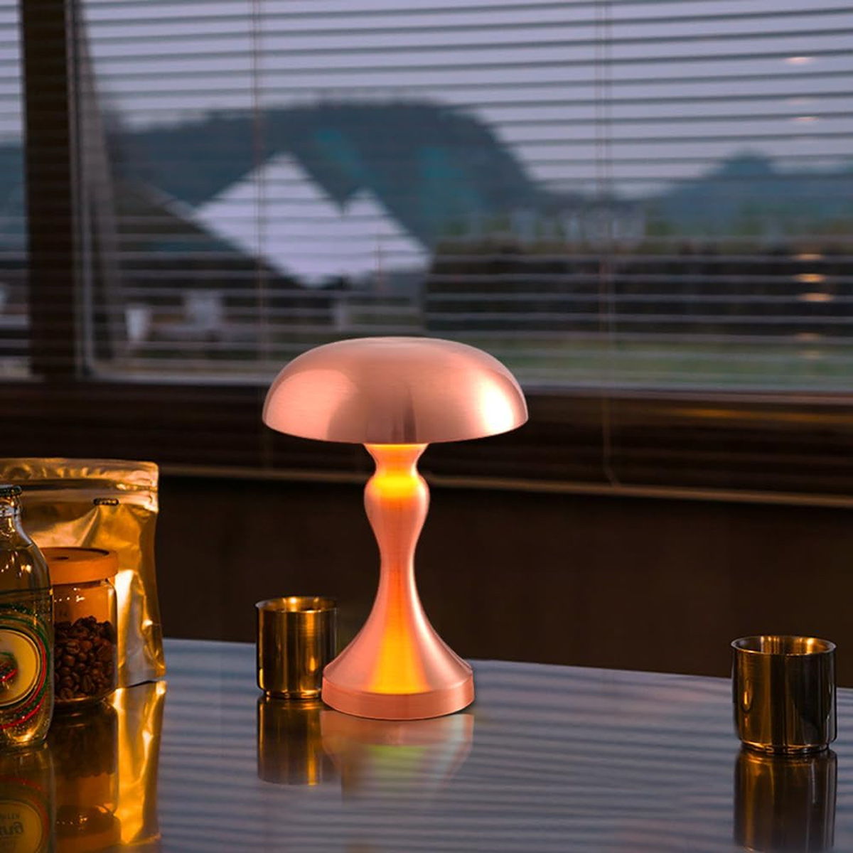 Auf einem Tisch steht eine Kupferlampe. Links sind Kaffeebohnen, ein Glas und eine Flasche. Im Hintergrund sind Jalousien und Berge.