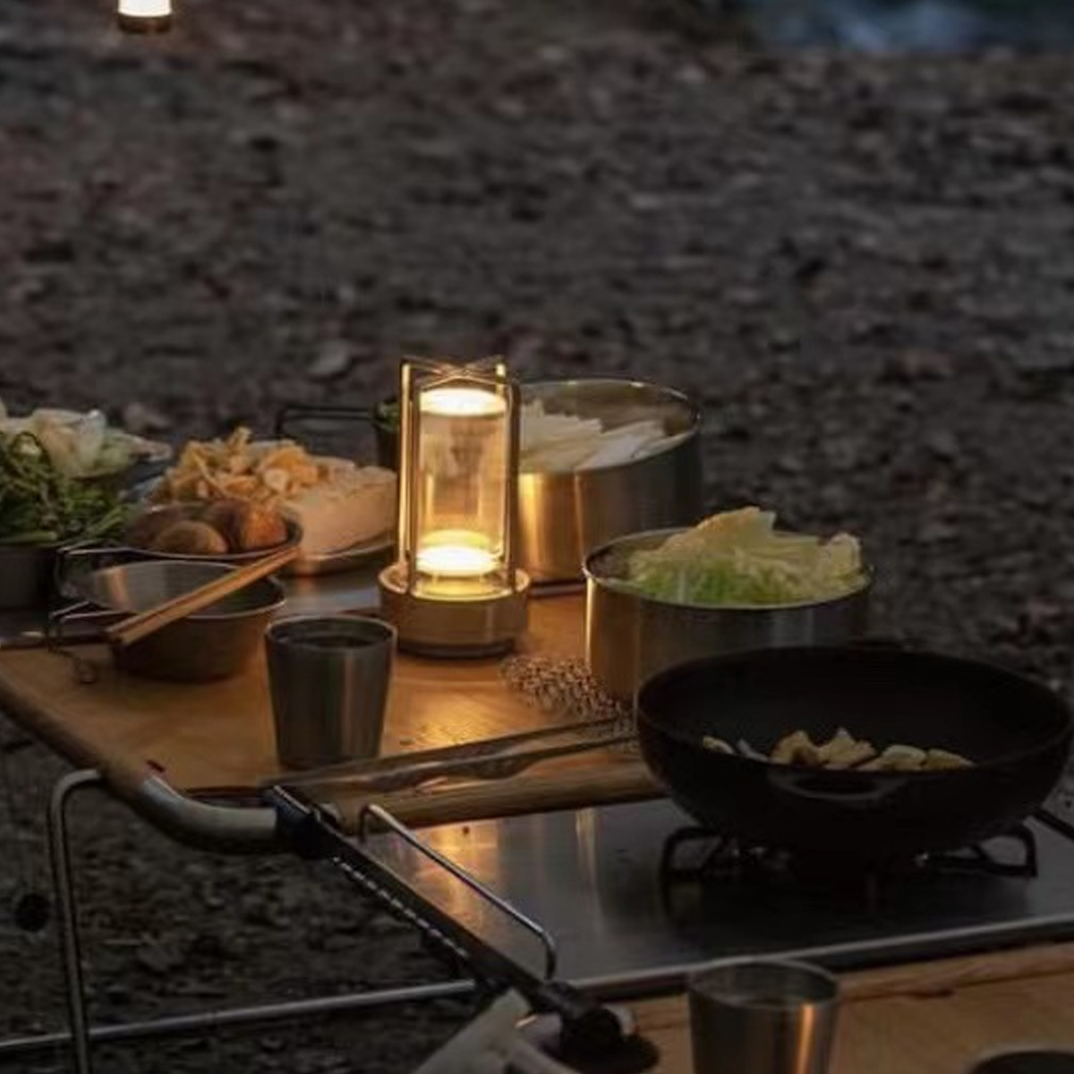 Ein Campingtisch mit Essen und einer Laterne. Gemüse, eine Pfanne mit Essen und Metallbecher sind vorhanden. Der Hintergrund ist eine verschwommene Naturkulisse.
