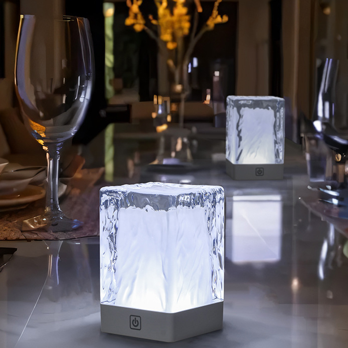 Drei beleuchtete, eiswürfelförmige Lampen auf einem Tisch. Weingläser und ein verschwommener Hintergrund mit Blumen sind ebenfalls zu sehen.