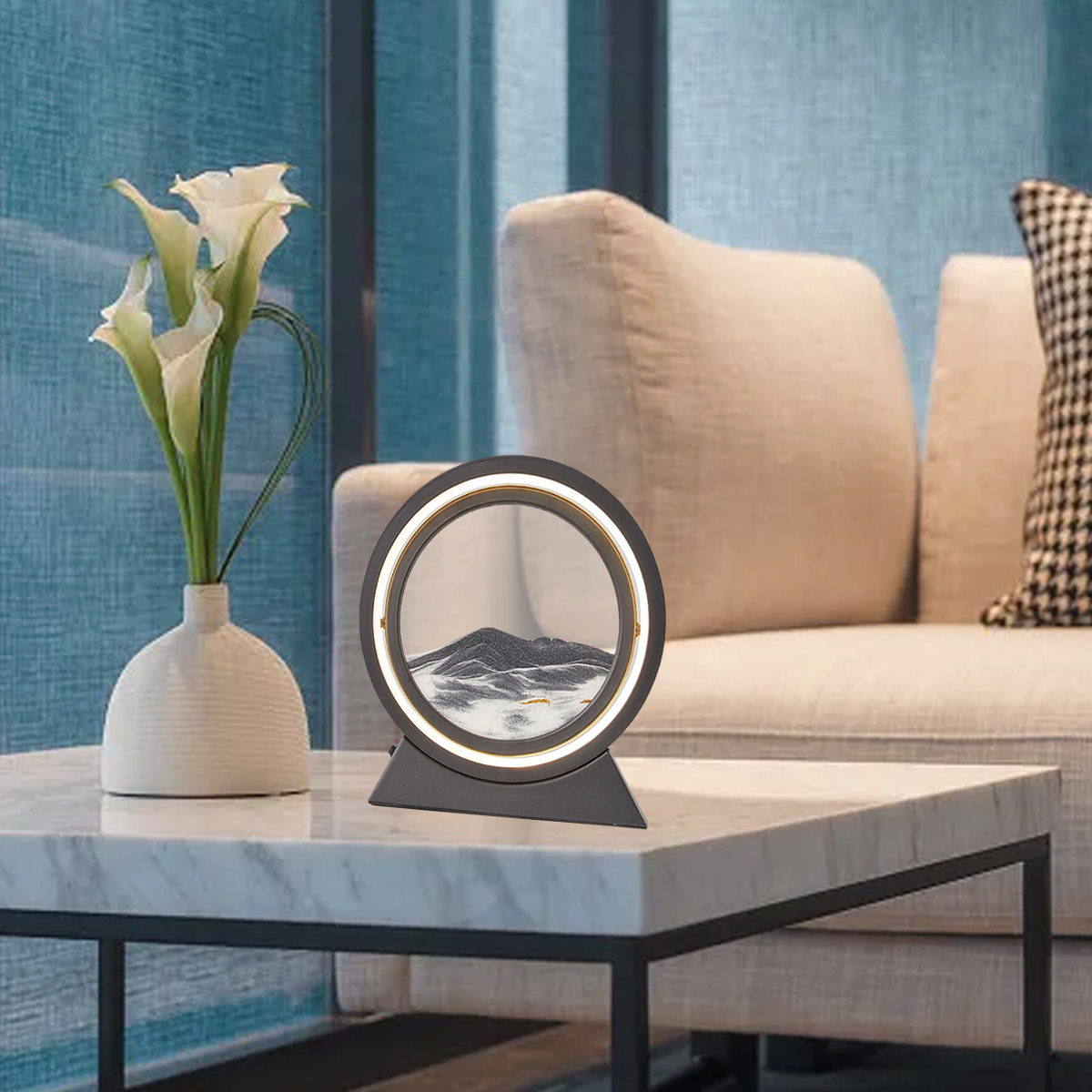 Auf einem Tisch stehen ein Sandkunstwerk, eine Vase mit Blumen und ein Sofa. Im Hintergrund befindet sich ein blauer Vorhang.