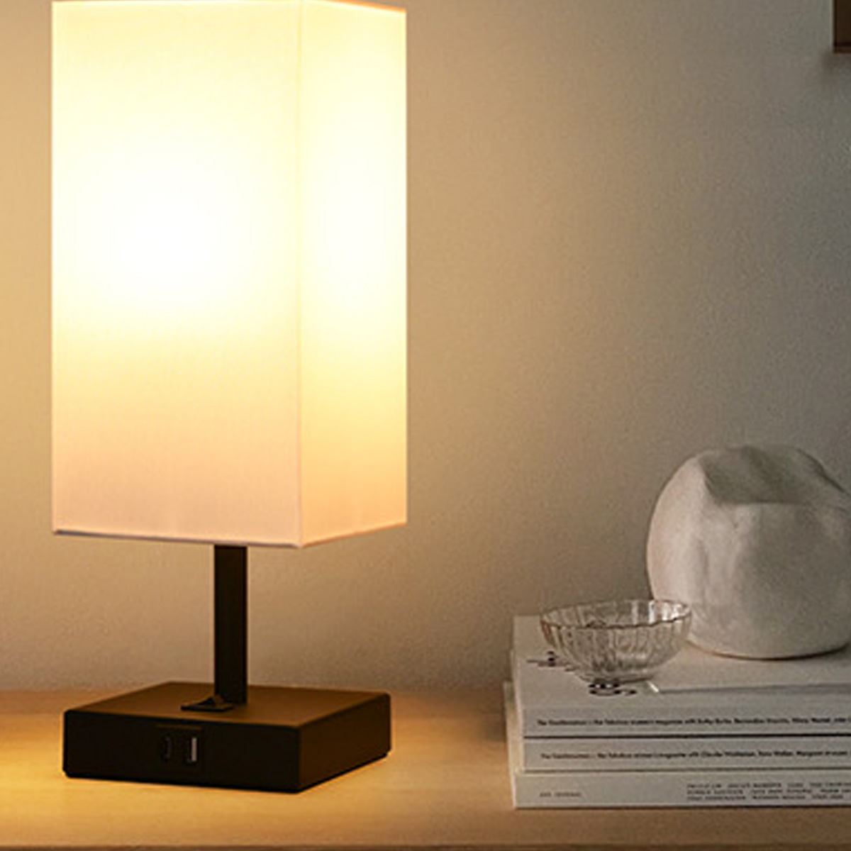 Eine beleuchtete rechteckige Lampe steht auf einem Tisch. Daneben Bücher, eine Glasschale und ein weißes Deko-Objekt. Warmes Licht geht aus.