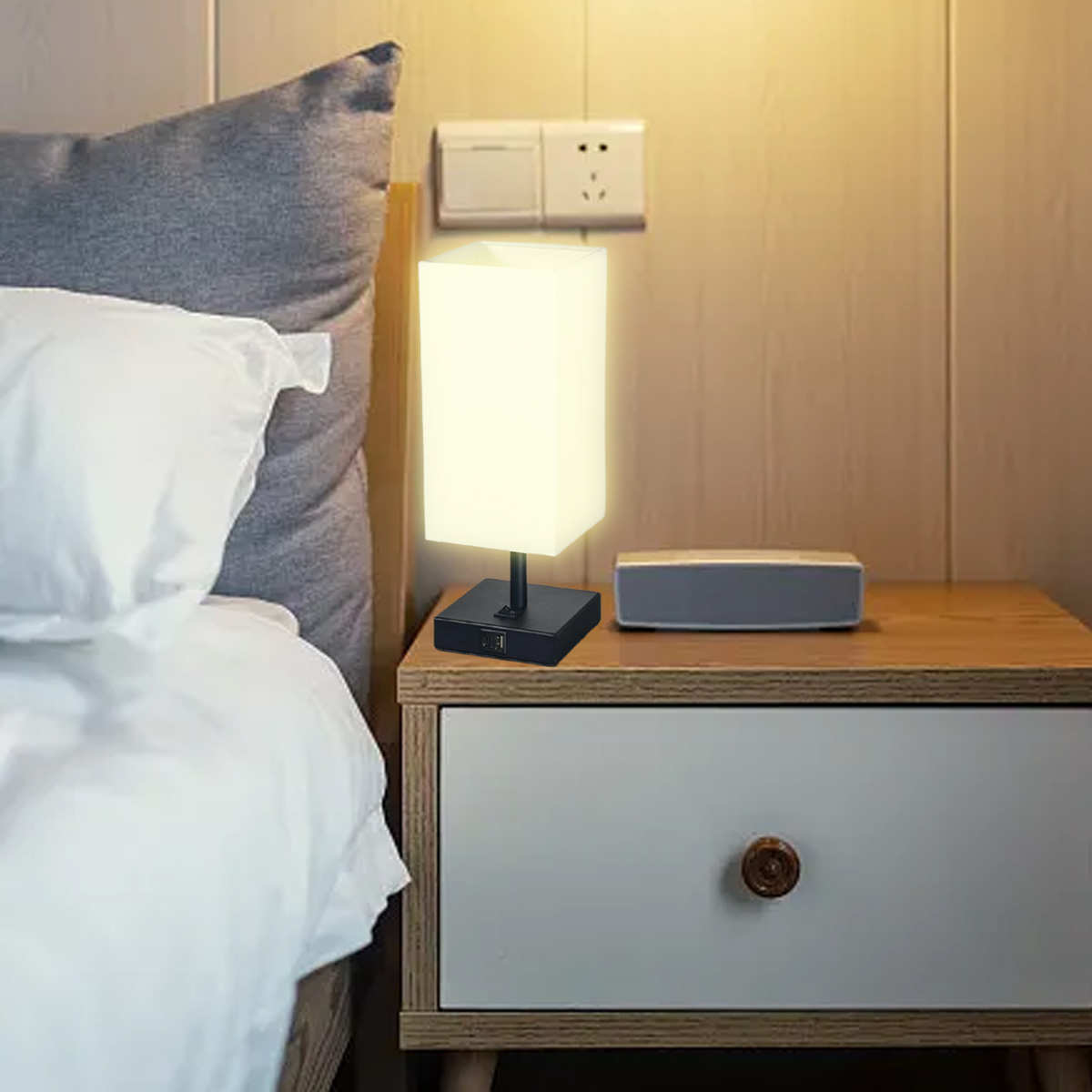 Eine beleuchtete Nachttischlampe steht auf einem Nachttisch. Links ist ein Bett. Ein Lautsprecher befindet sich neben der Lampe.