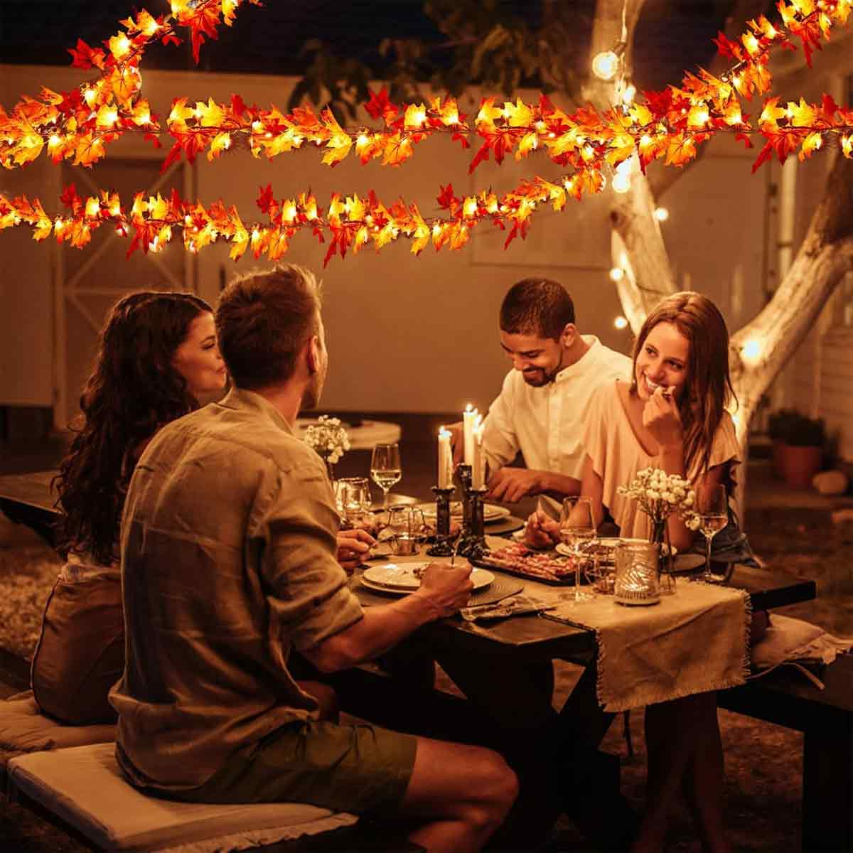Vier Personen sitzen draußen an einem Esstisch. Herbstblätter und Lichter hängen darüber. Kerzen und Essen stehen auf dem Tisch.