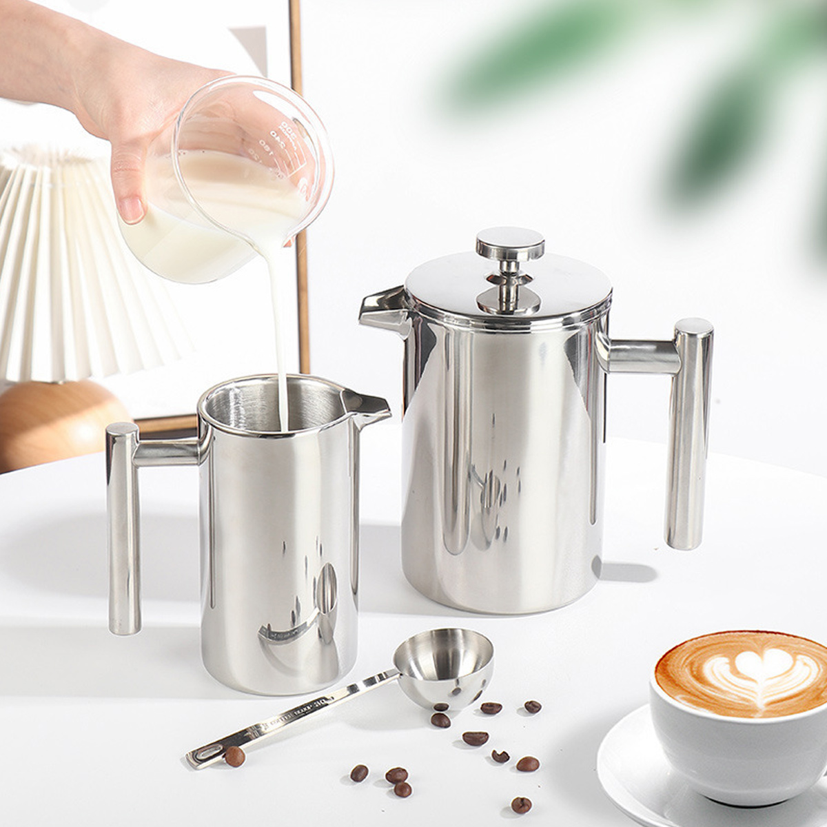 Eine Hand gießt Milch aus einem Becher in einen Edelstahlkrug. Eine French Press, ein Löffel, Kaffee und Bohnen liegen auf einer weißen Oberfläche.