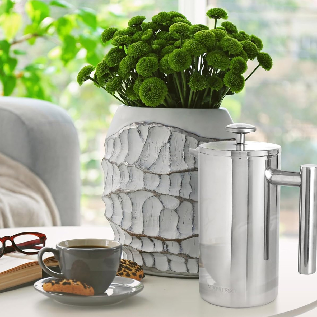 Kaffee-Setup mit French Press, Blumen, Kaffeetasse und Keksen auf einem weißen Tisch am Fenster mit Grün.