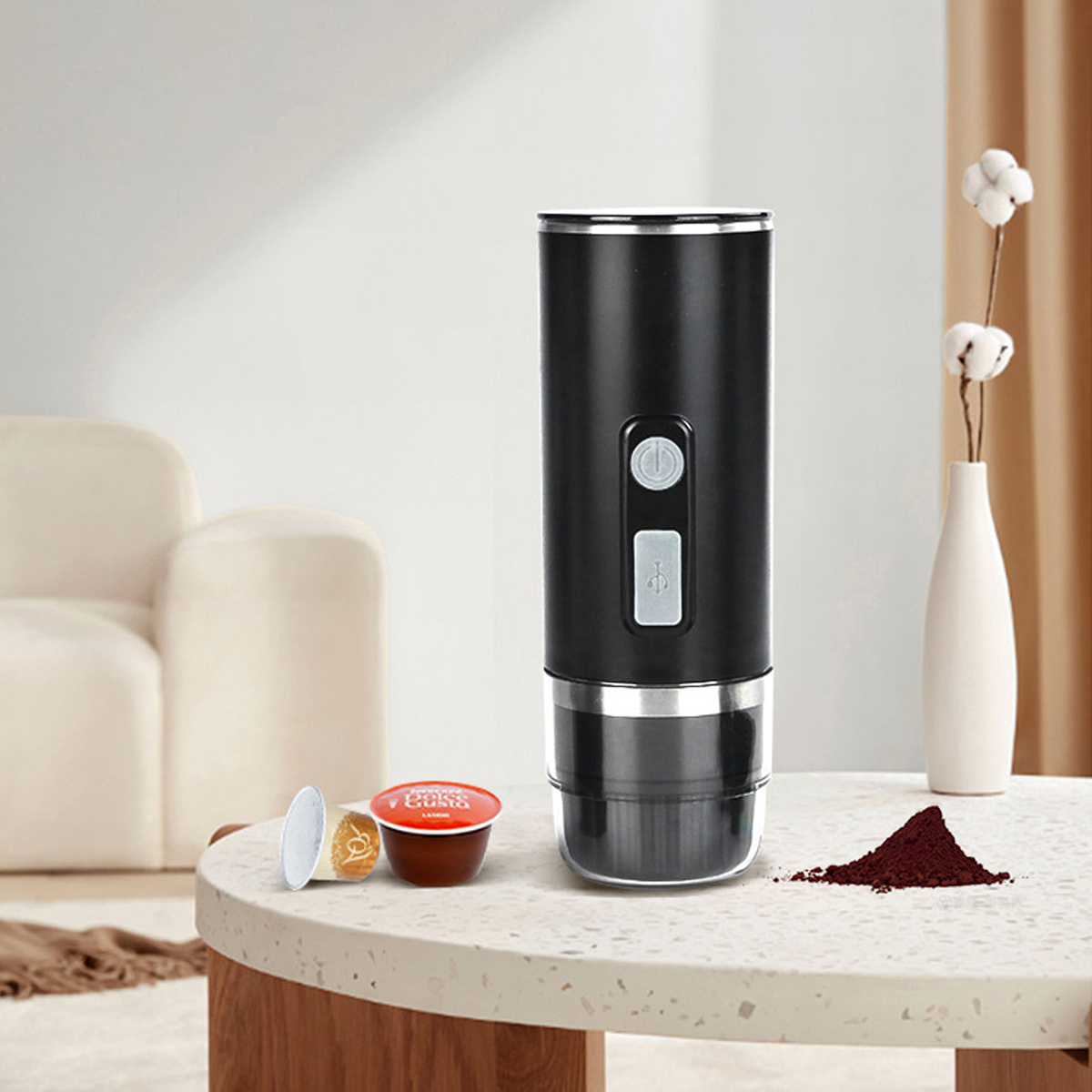Ein schwarzer Kaffeezubereiter mit Kaffeekapseln steht auf einem Tisch. Kaffeepulver und eine Vase mit Baumwollblüten sind ebenfalls vorhanden.