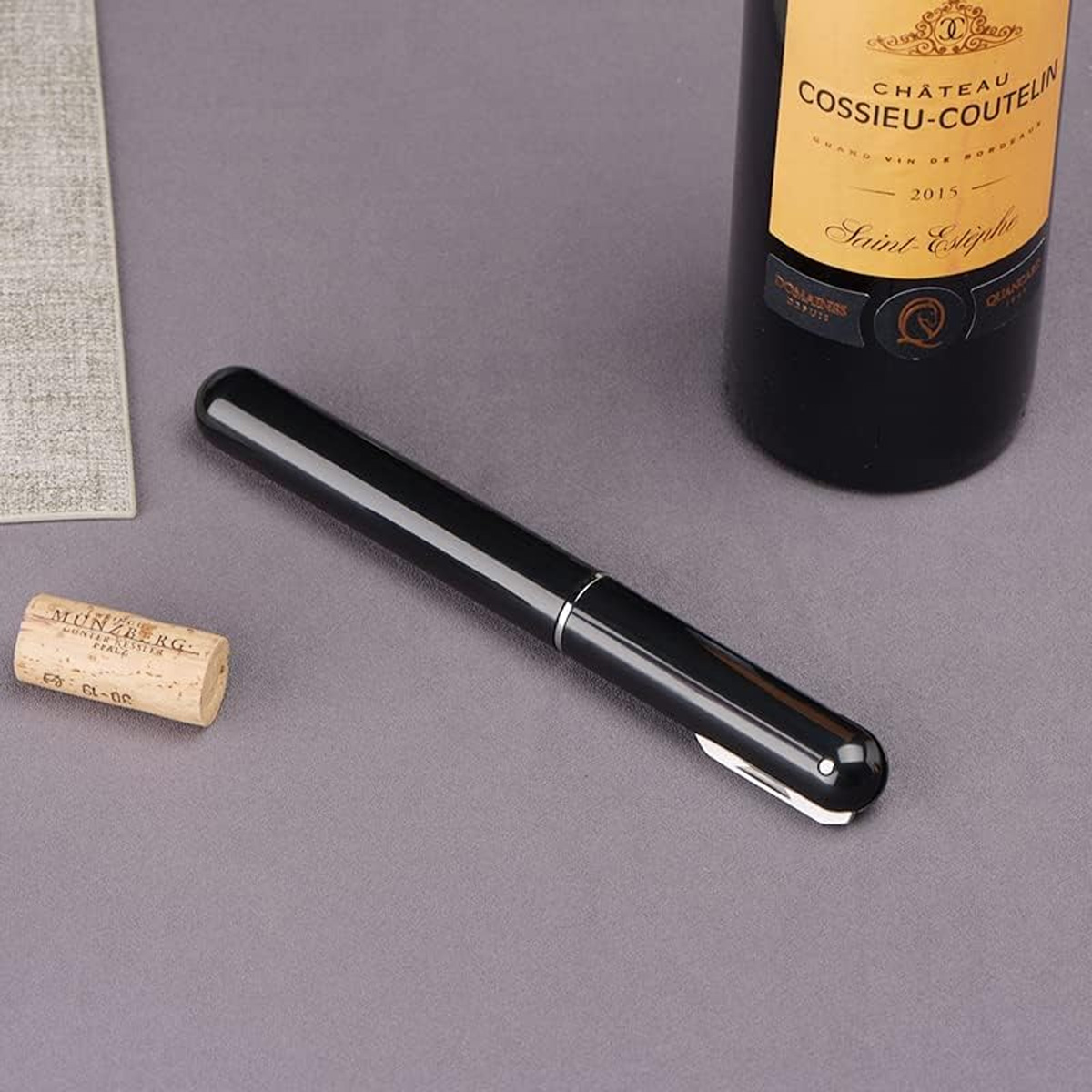 Ein schwarzer Stift liegt auf einer grauen Oberfläche neben einer Weinflasche und einem Korken. Die Flasche hat ein Etikett und der Korken Text.