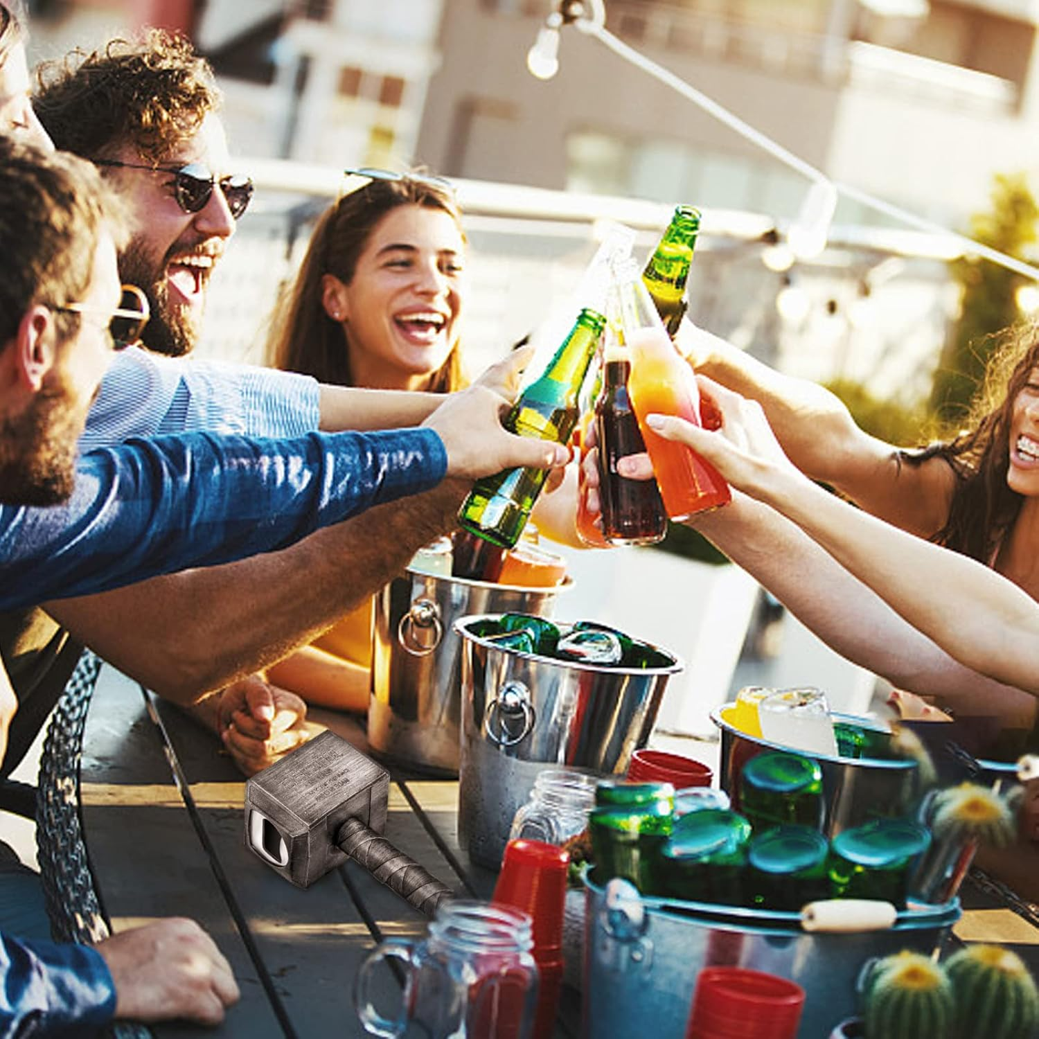 Eine Gruppe von Freunden stoßt mit Getränken auf einer Dachterrasse an. Bierflaschen und Cocktails werden hochgehalten. Ein Thor-Hammer liegt auf dem Tisch.