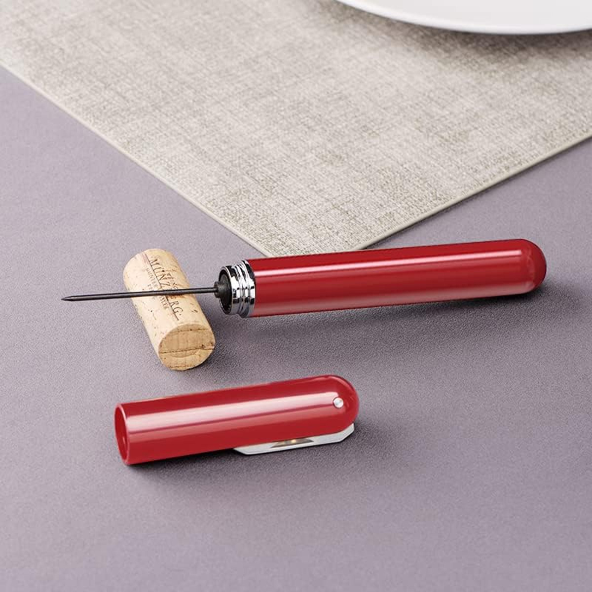 Ein roter Weinöffner mit Korken und Abdeckung auf grauem Untergrund, mit Tischset und weißem Teller im Hintergrund.