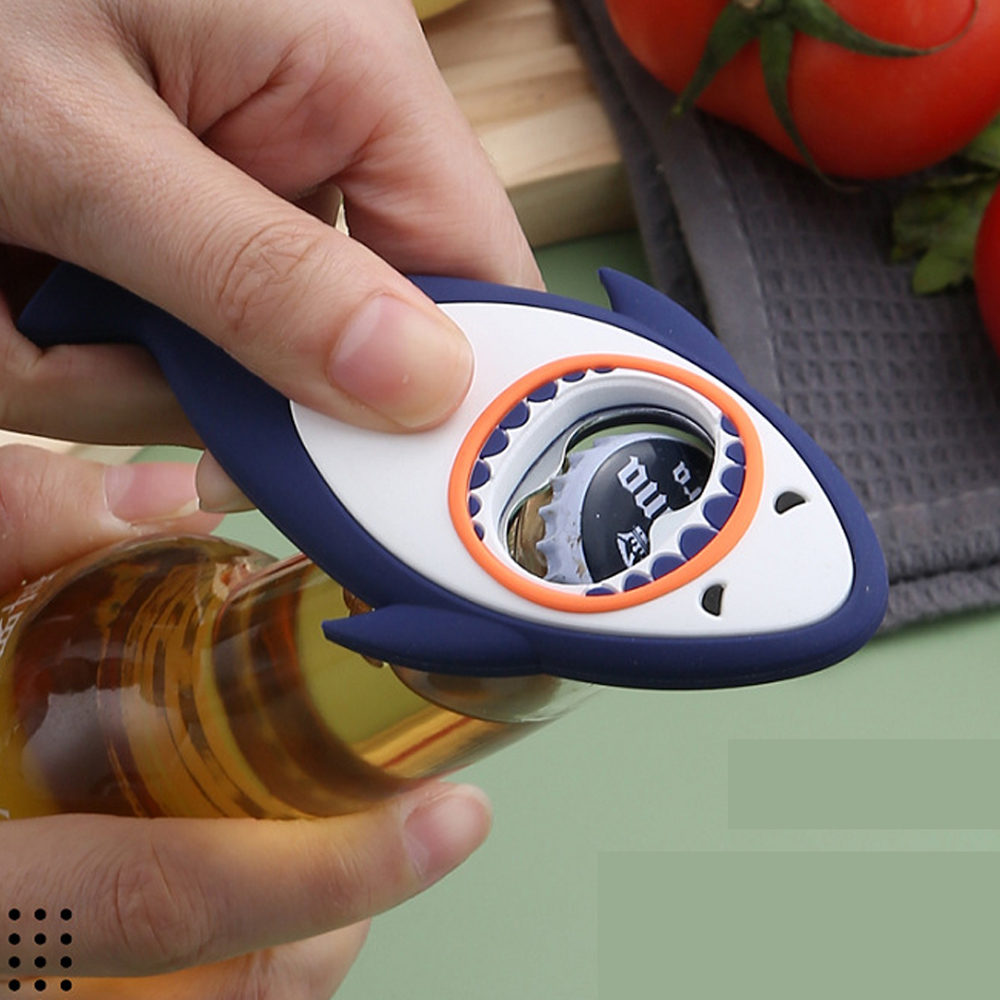 Ein haiförmiger Flaschenöffner wird verwendet, um eine Bierflasche zu öffnen. Hände halten den Öffner, Tomaten und Stoff sind im Hintergrund.