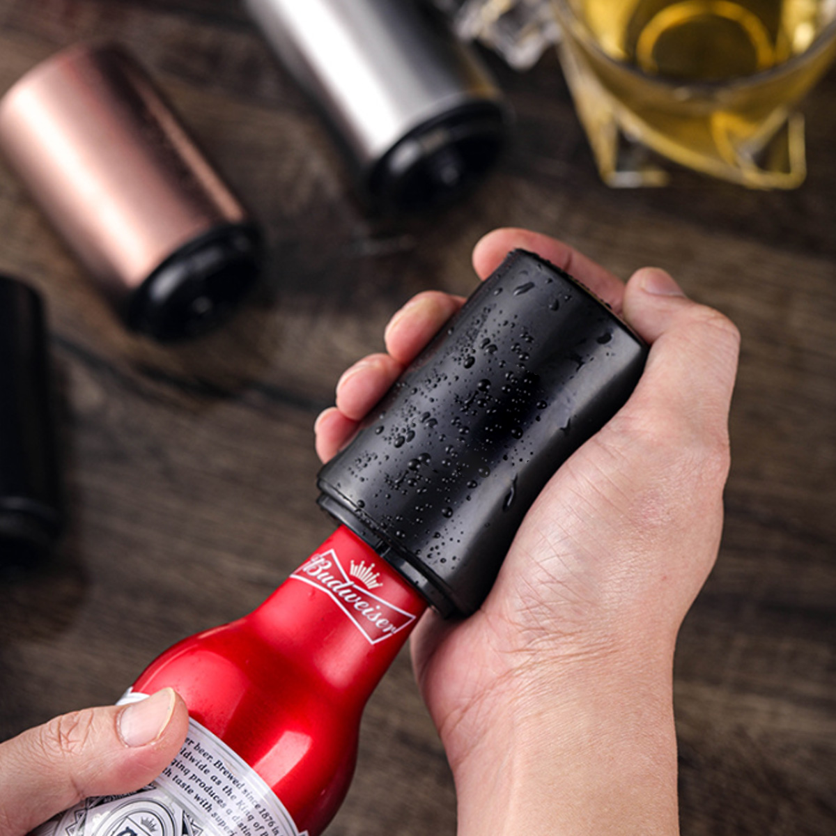 Eine Hand hält einen schwarzen Flaschenöffner über einer roten Budweiser-Flasche. Andere Öffner und ein Bierglas liegen auf Holz.