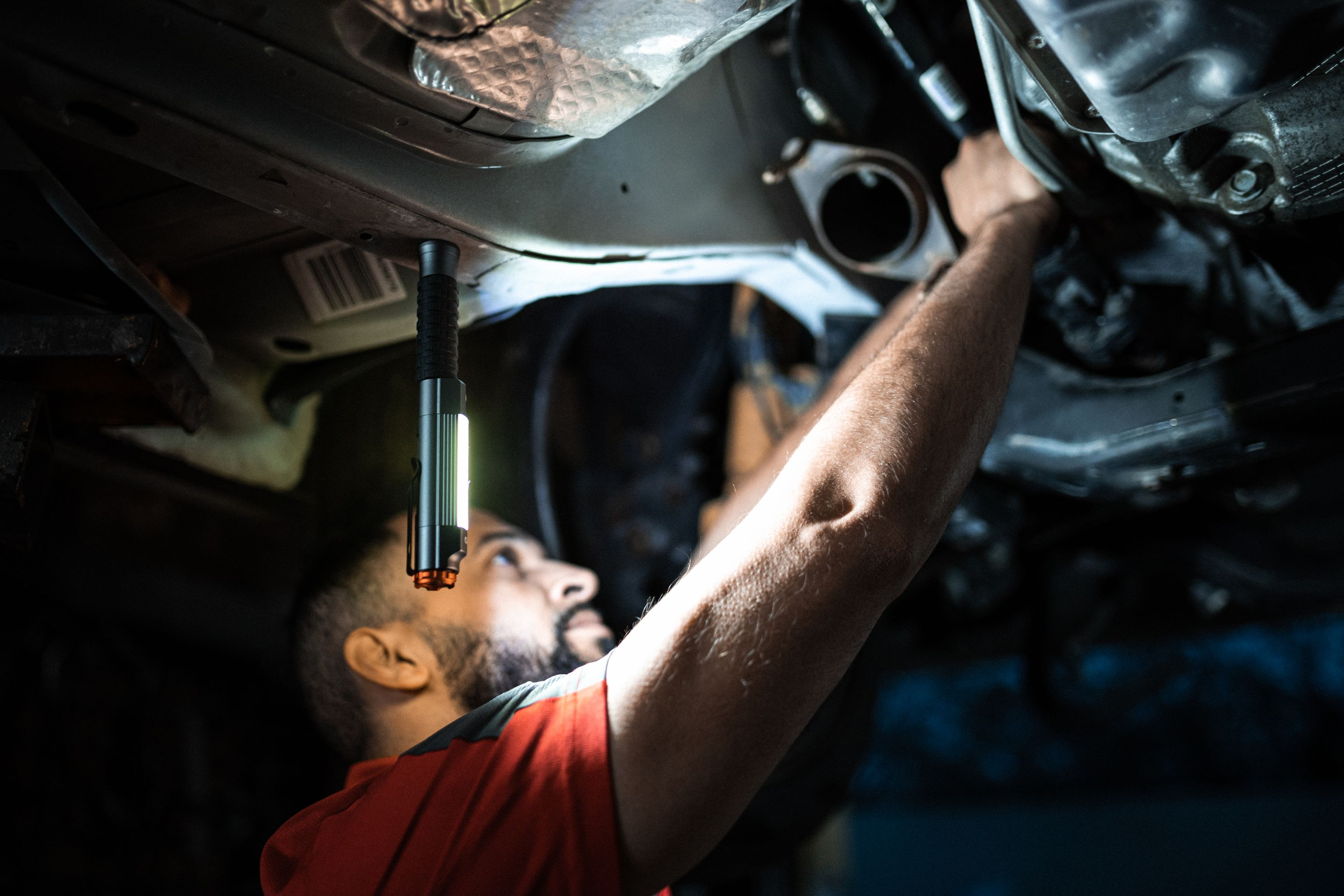 Ein Mechaniker arbeitet unter einem Auto, beleuchtet von einer Arbeitsleuchte. Er trägt ein rotes Hemd und schaut nach oben.