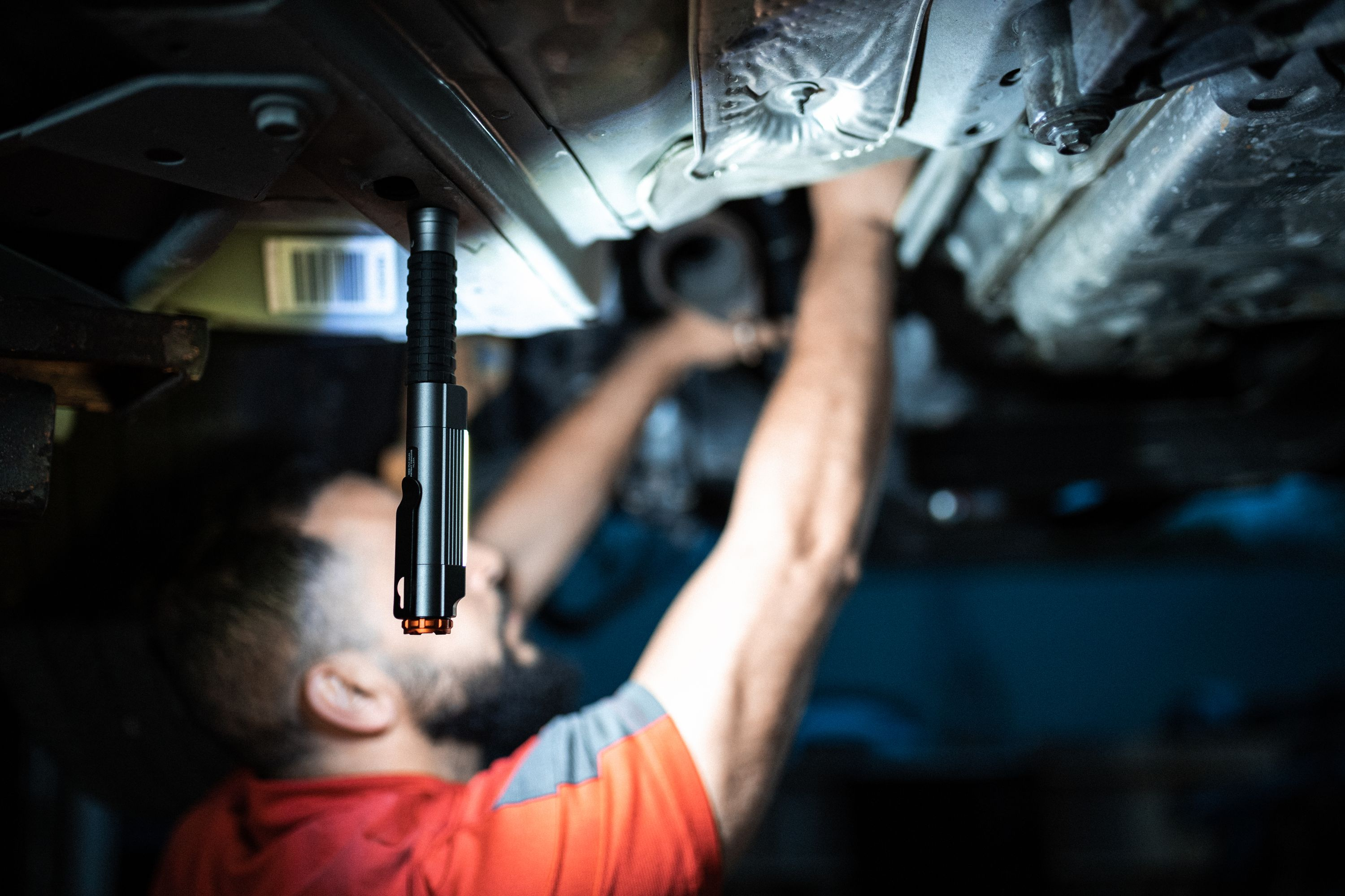 Ein Mechaniker arbeitet unter einem Auto, beleuchtet von einer Taschenlampe. Der Mechaniker trägt ein rotes Hemd.