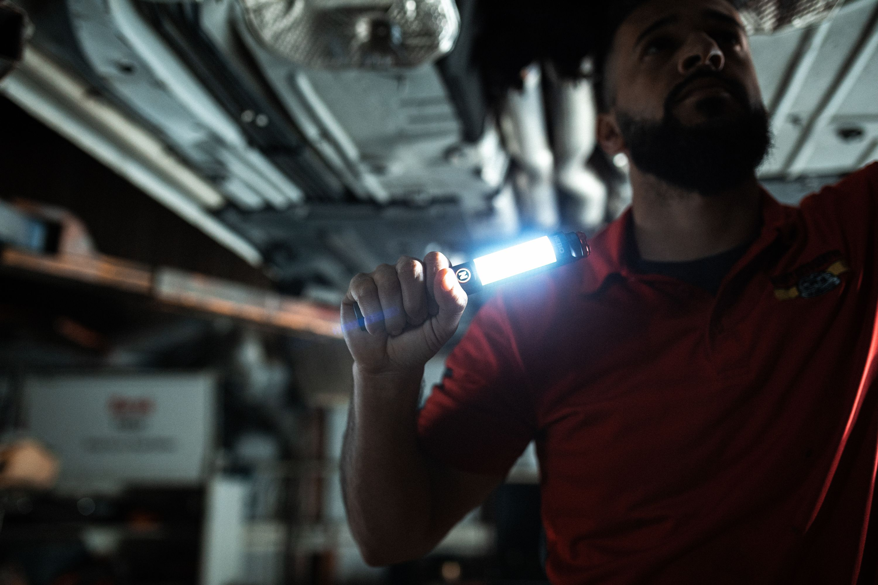Mechaniker unter einem Auto, der eine helle Lampe hält. Er trägt ein rotes Hemd. Die Unterseite des Autos ist sichtbar.