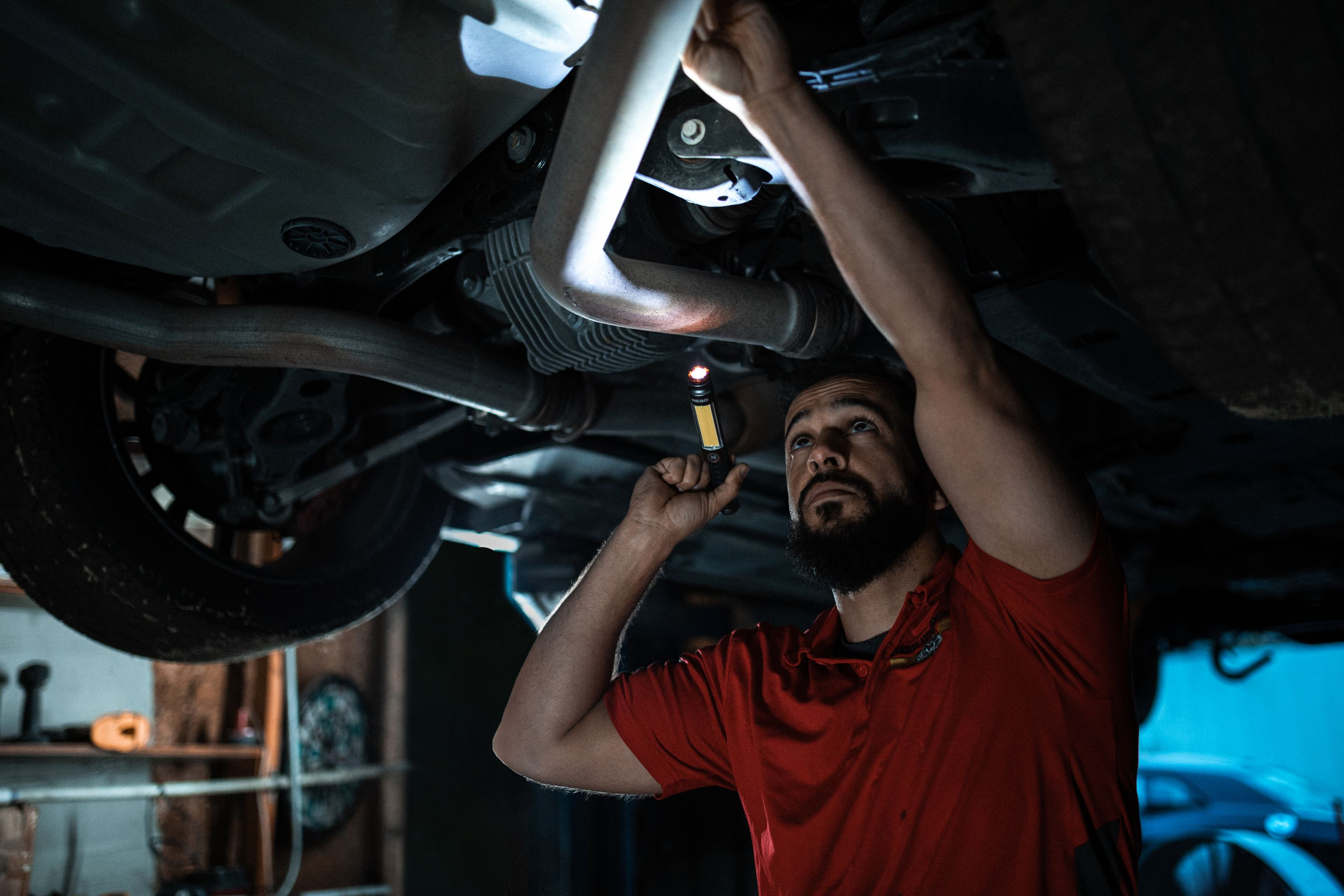 Mechaniker unter dem Auto, hält eine Taschenlampe. Rotes Hemd, Bart. Autoteile über Kopf. Dunkle Umgebung, wahrscheinlich eine Garage.