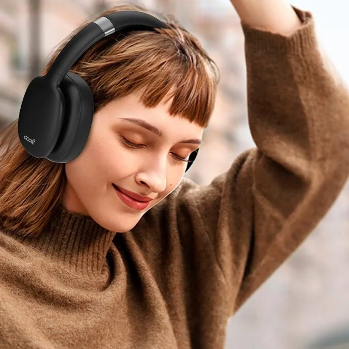 Una mujer con auriculares negros sonríe. Tiene cabello castaño y un suéter marrón. Tiene el brazo levantado.
