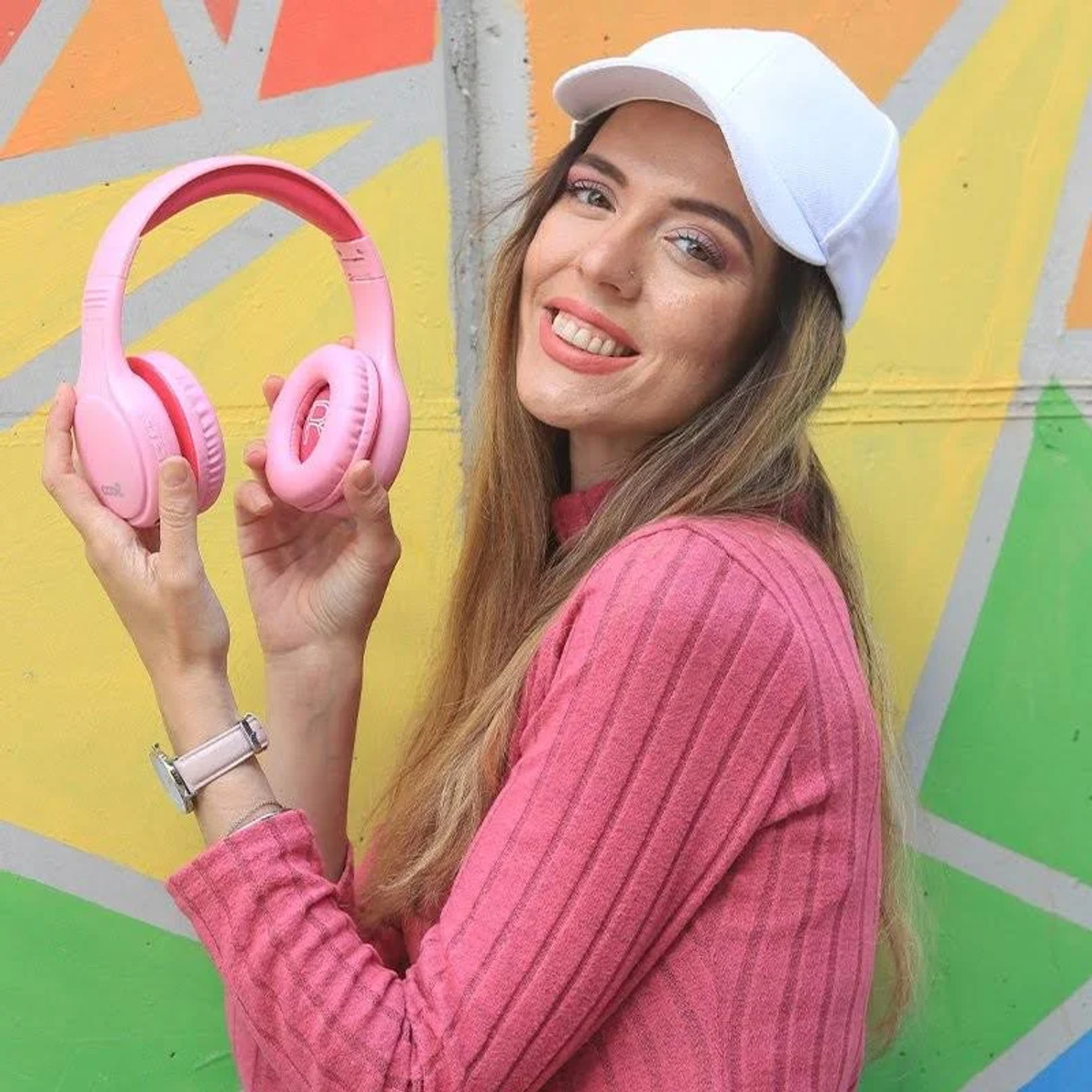 Mujer con gorra blanca, suéter rosa y auriculares sonríe. Pared colorida detrás.