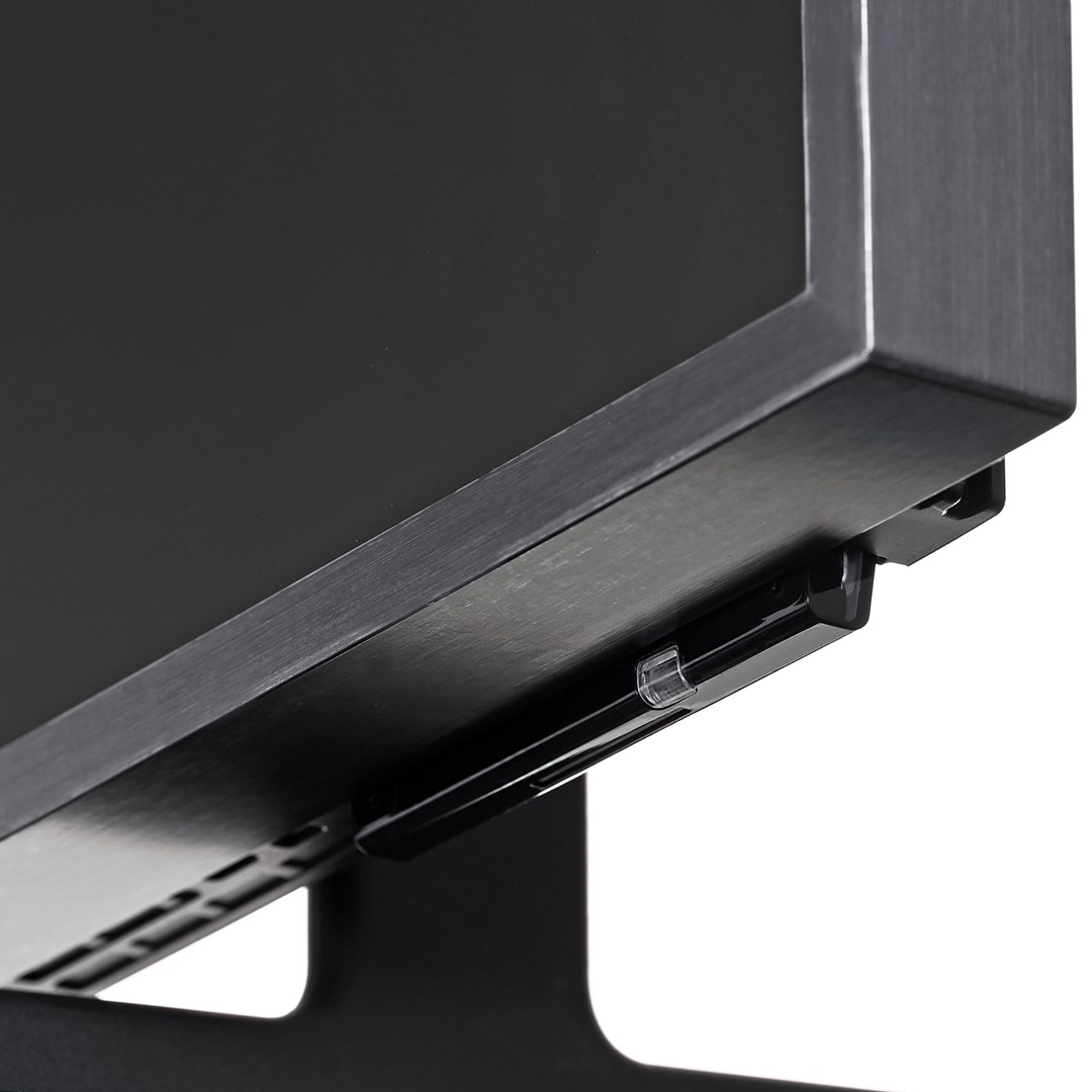 Close-up van de onderrand van een zwarte monitor, met een zwarte voet. Een zilveren frame omgeeft het scherm. Een zwart, rechthoekig apparaat is bevestigd.