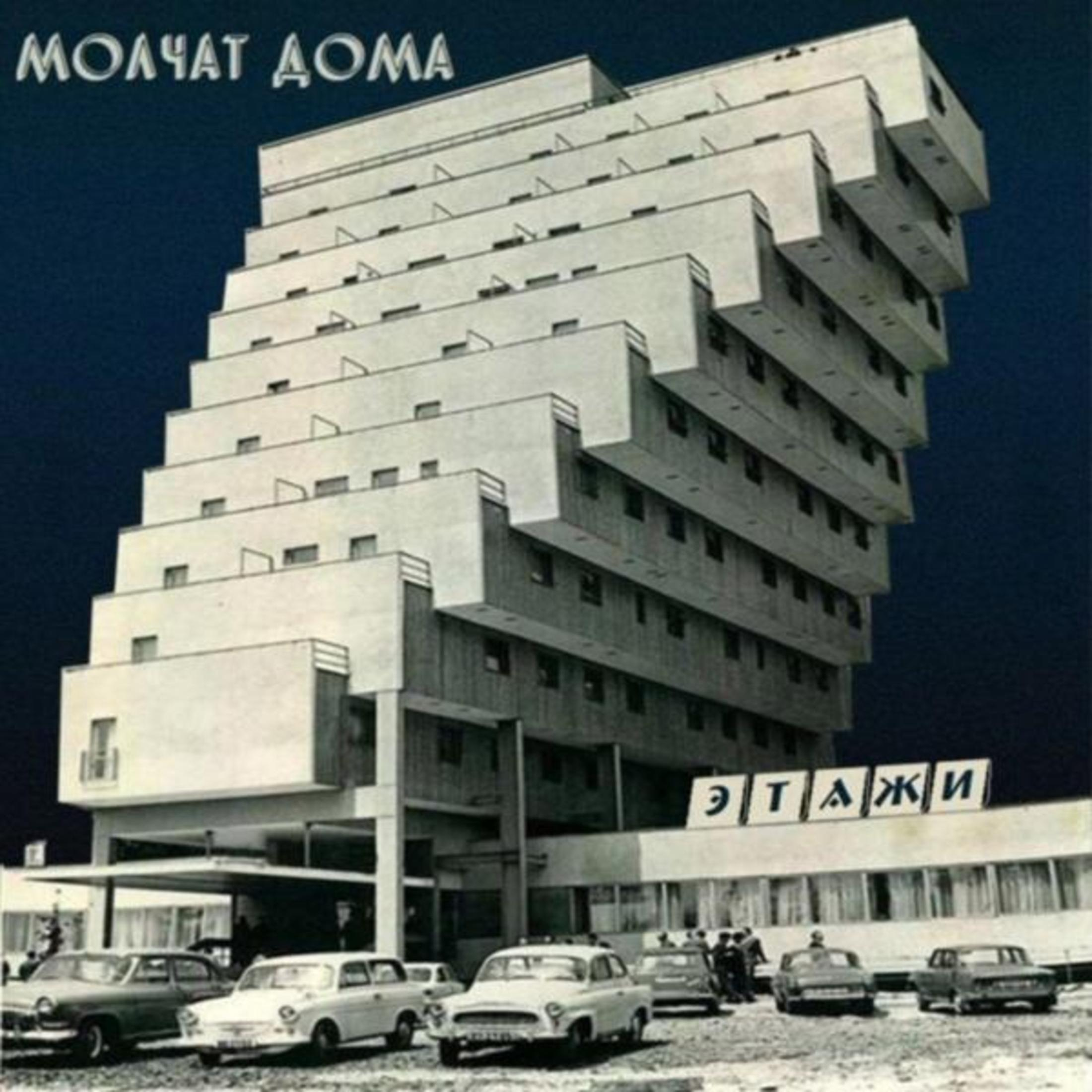 Un edificio bianco angolato con molti livelli. Ci sono auto davanti. Il cielo è blu scuro. In alto c'è scritto 'MOLCHAT DOMA'.