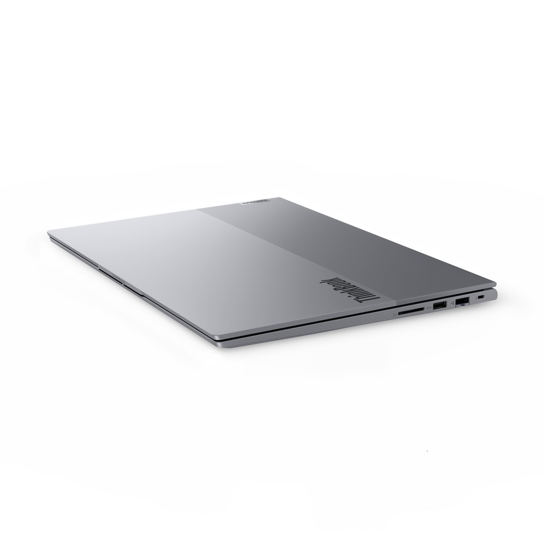 Szary laptop ThinkBook jest lekko otwarty, ustawiony pod kątem na białej powierzchni. Widoczne jest logo Lenovo.