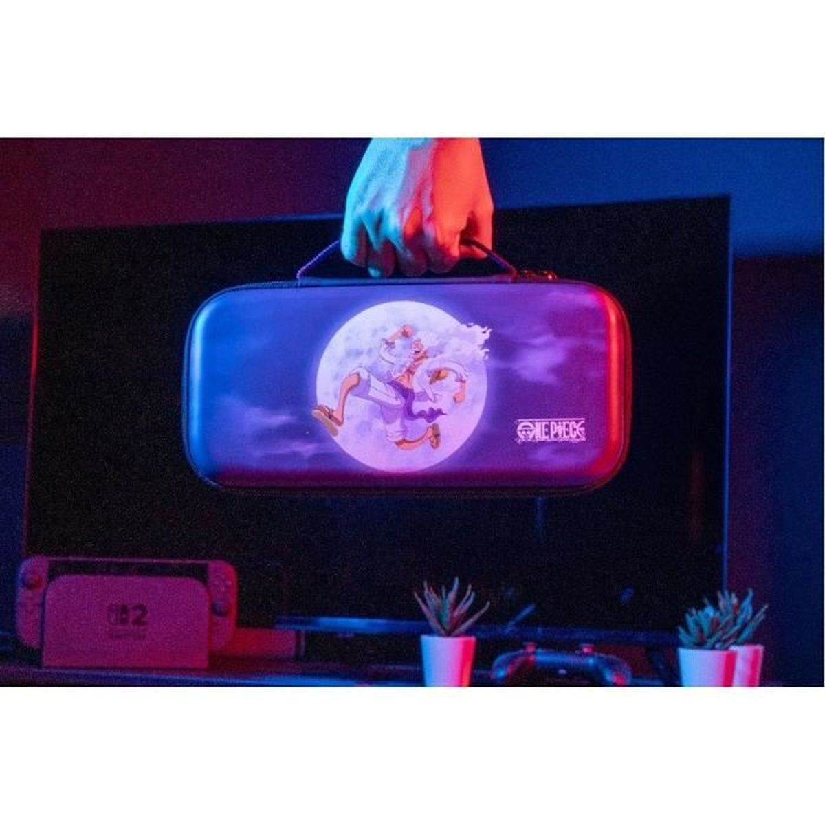 A hand holds a One Piece themed case featuring a character in front of a moon, with a Nintendo Switch and plants in the background.