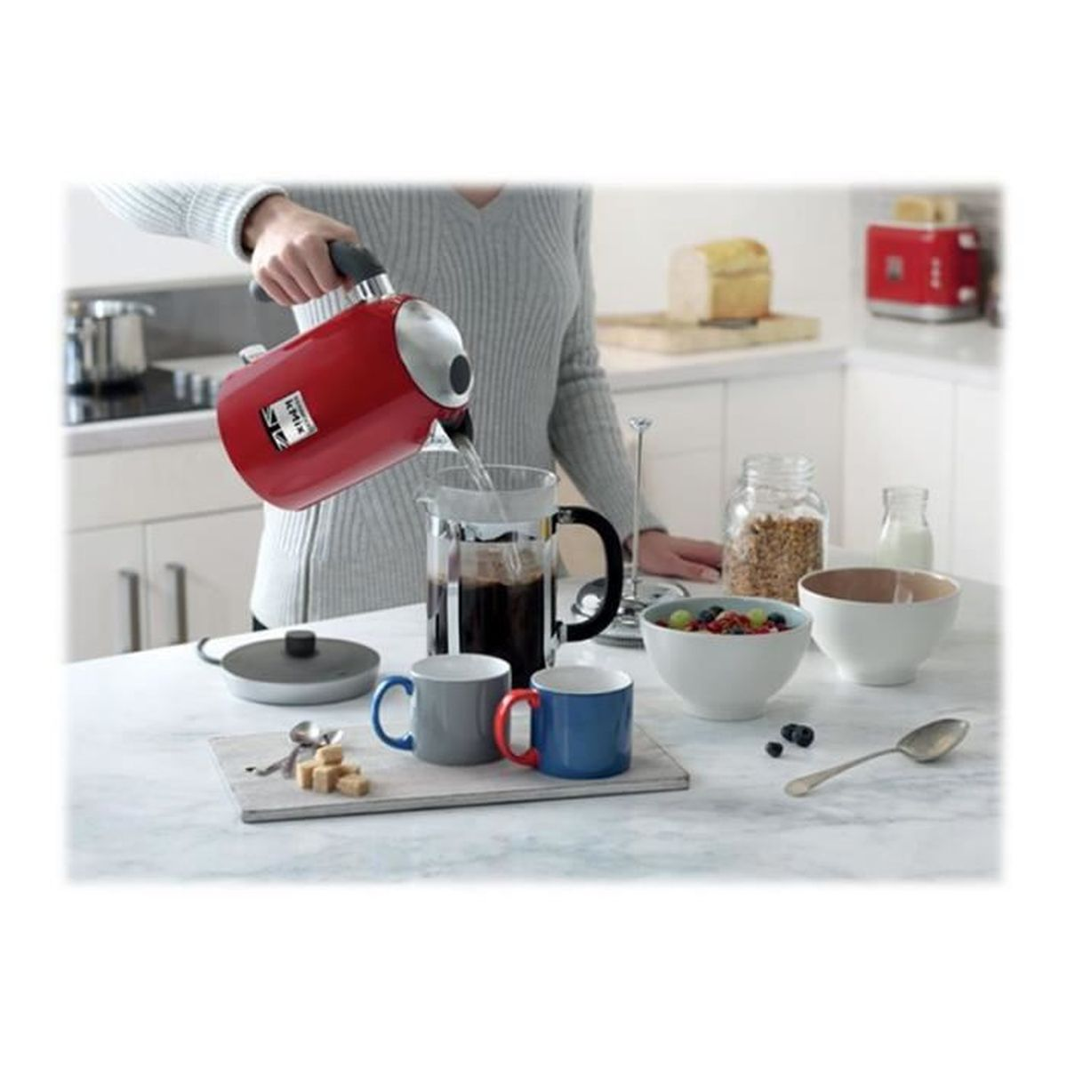 Osoba wlewa wodę z czerwonego czajnika do ekspresu do kawy. Na białej powierzchni znajdują się kubki, miski i toster.