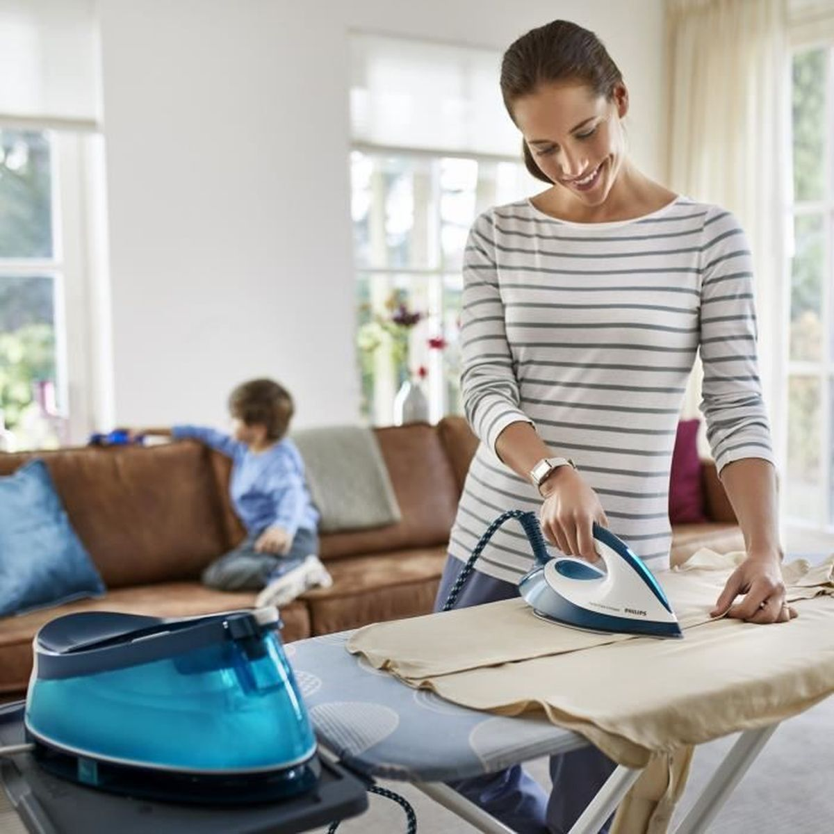 Kobieta prasuje ubrania. Dziecko siedzi na kanapie w tle. Widoczne żelazko i niebieski zbiornik na wodę.