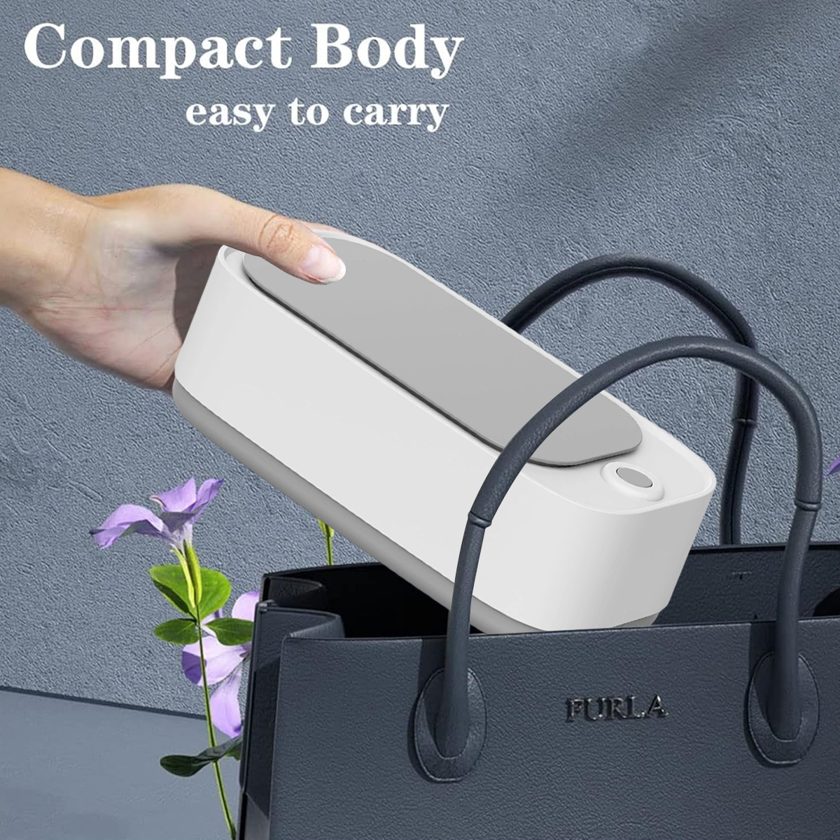 Eine Hand legt ein weißes und graues Gerät in eine dunkelgraue Furla-Tasche. Lila Blumen befinden sich in der linken unteren Ecke, mit Text oben.