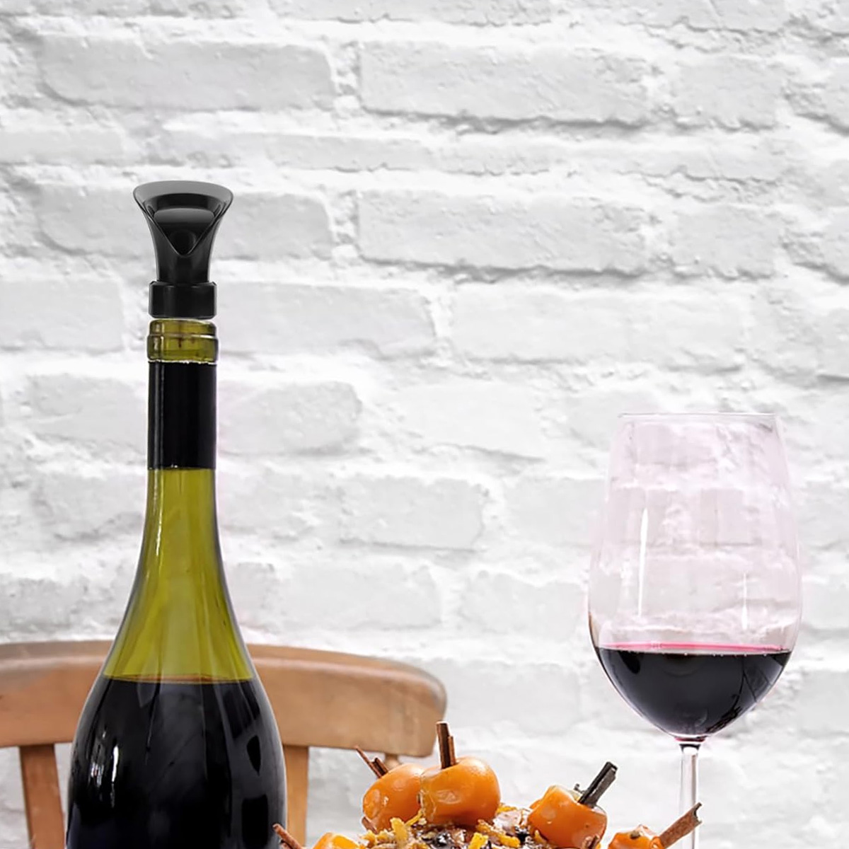 Eine Weinflasche mit einem Weinbelüfter, ein Glas Rotwein und ein dekoratives Gericht auf einem Tisch mit weißer Backsteinmauer.