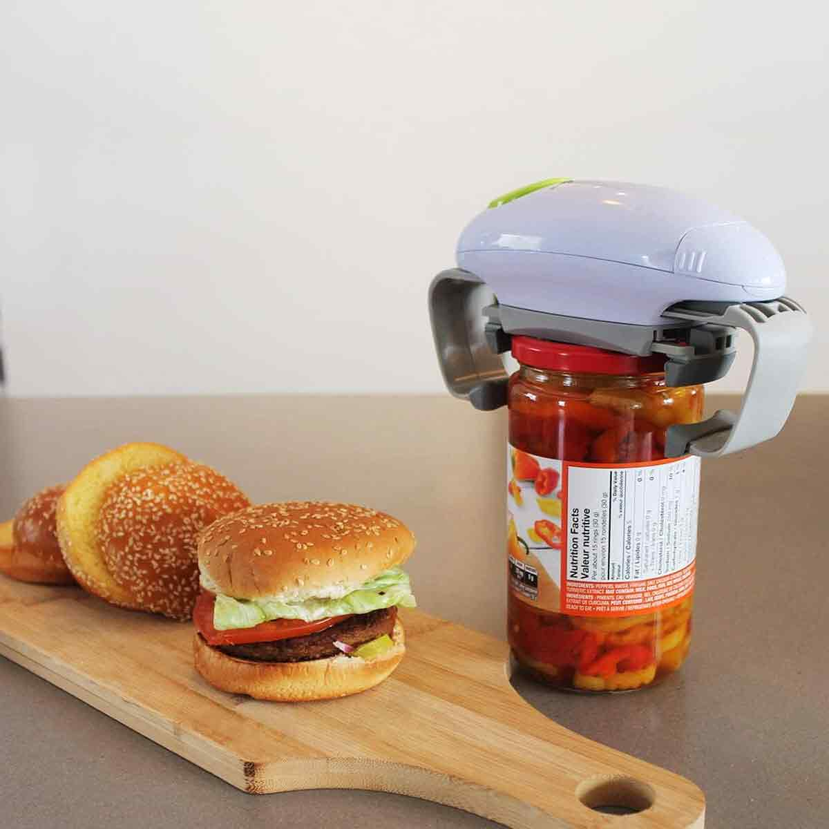 Ein Hamburger und Brötchen auf einem Holzbrett. Ein Glas mit einem Glasöffner. Der Hintergrund ist eine weiße Wand. Das Glas hat rote Paprika.