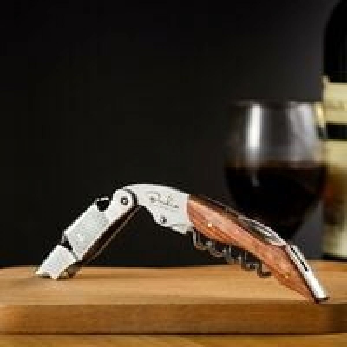Ein Weinöffner auf einer Holzoberfläche. Ein Weinglas, eine Weinflasche und ein dunkler Hintergrund sind sichtbar.